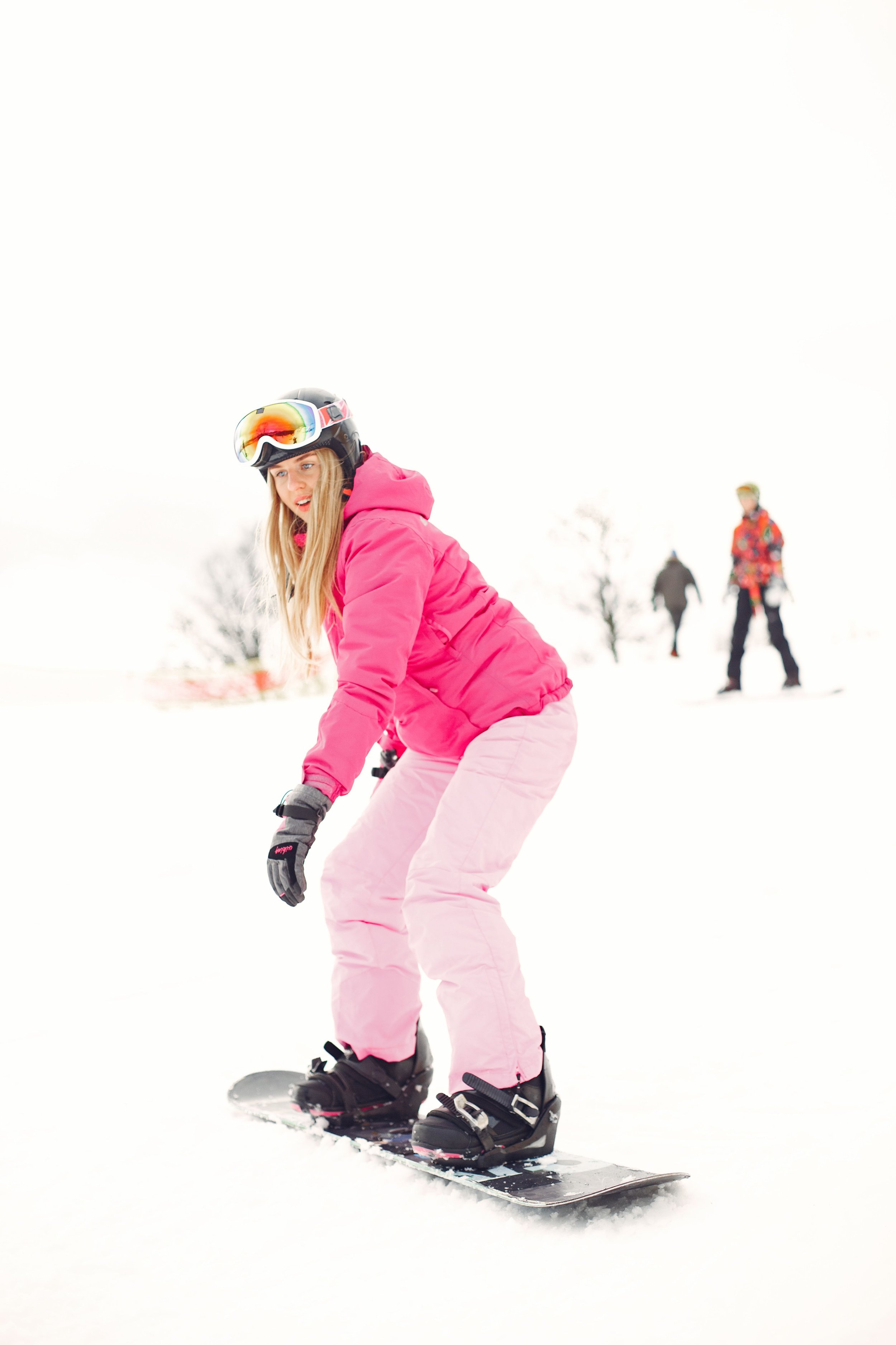 A young girl in a pink jacket and snow pants snowboarding in a snowy landscape with two other people in the background.
