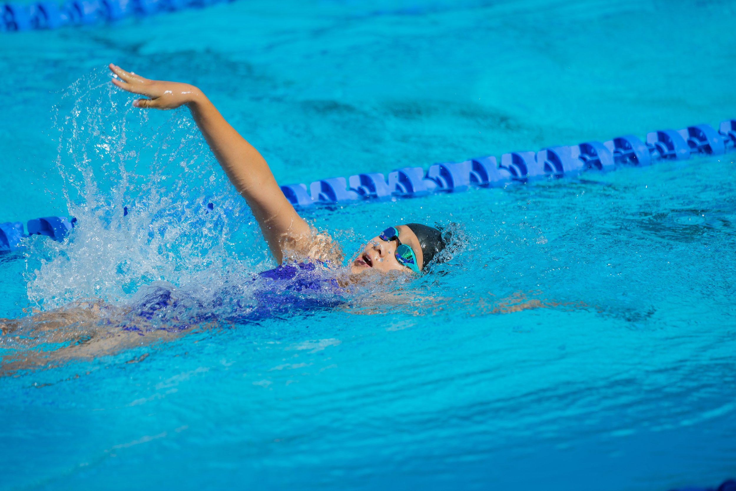 A young swimmer wearing goggles and a swim cap, swimming freestyle in a blue swimming pool with lane markers.