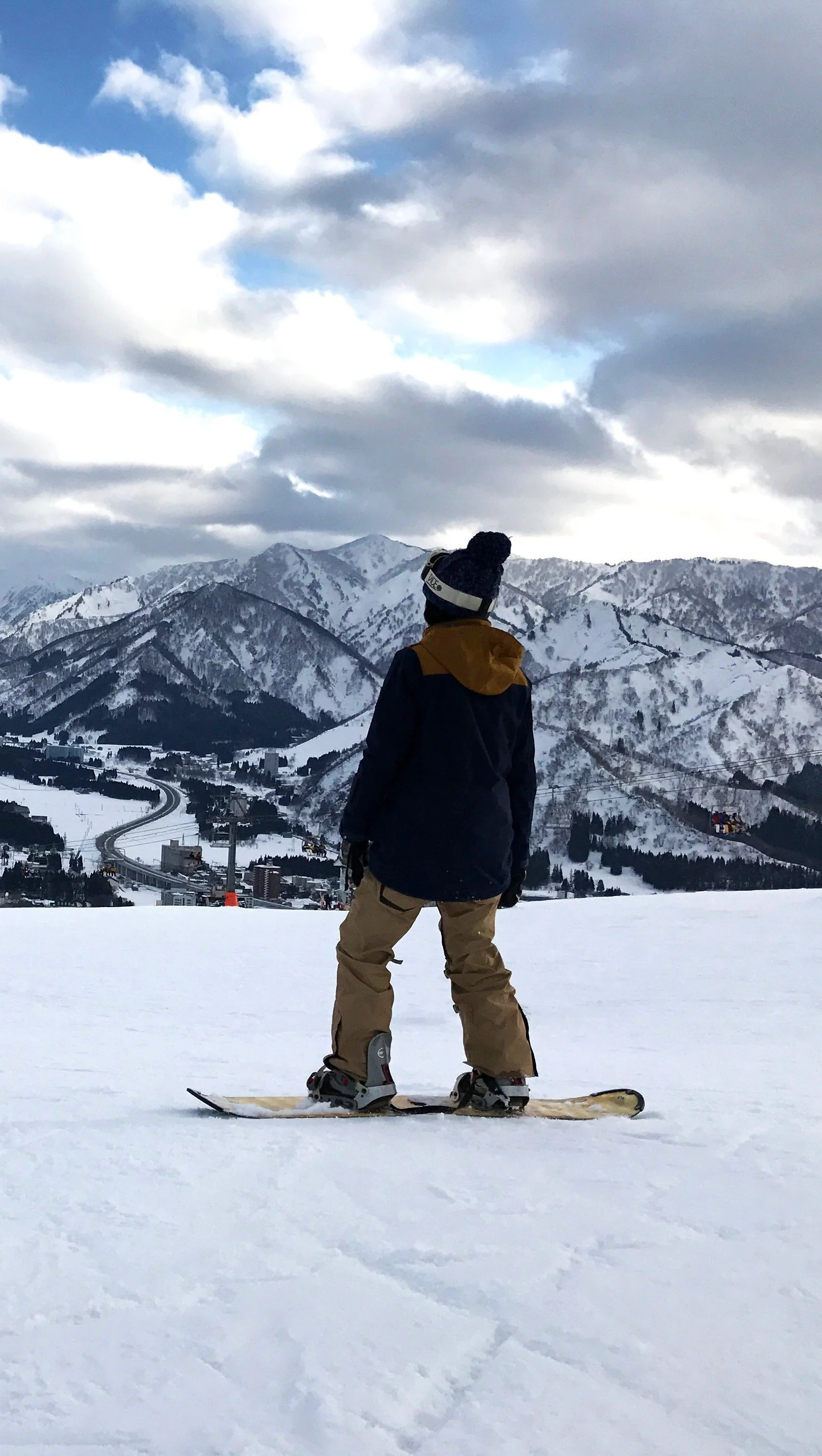 A person standing on a snowboard in a snowy mountain landscape, wearing winter gear and looking at the snow-covered mountains and cloudy sky.