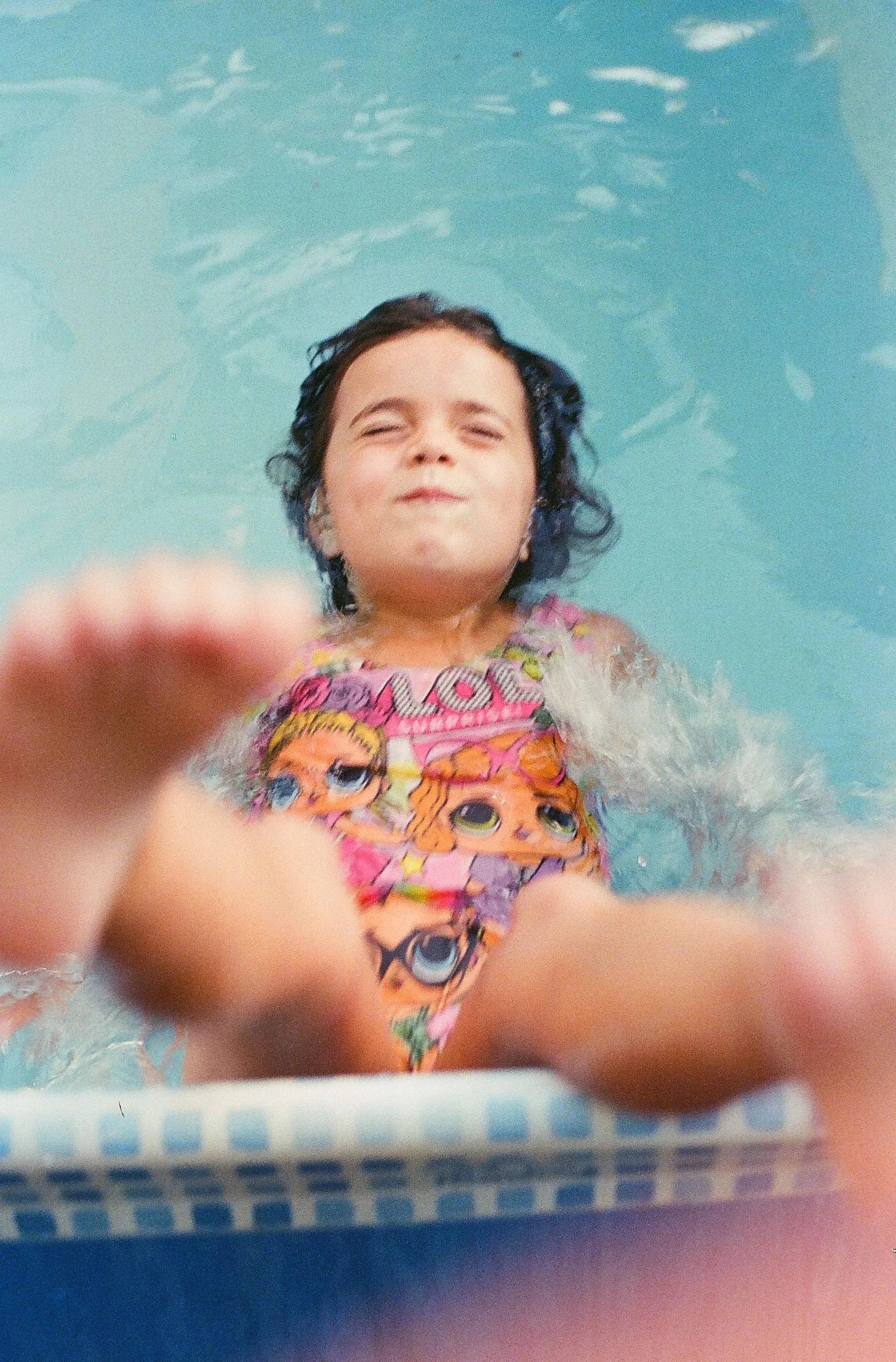 Young girl with wet hair floating in a swimming pool, wearing a colorful swimsuit with cartoon characters, smiling with eyes closed, fingers near the camera.