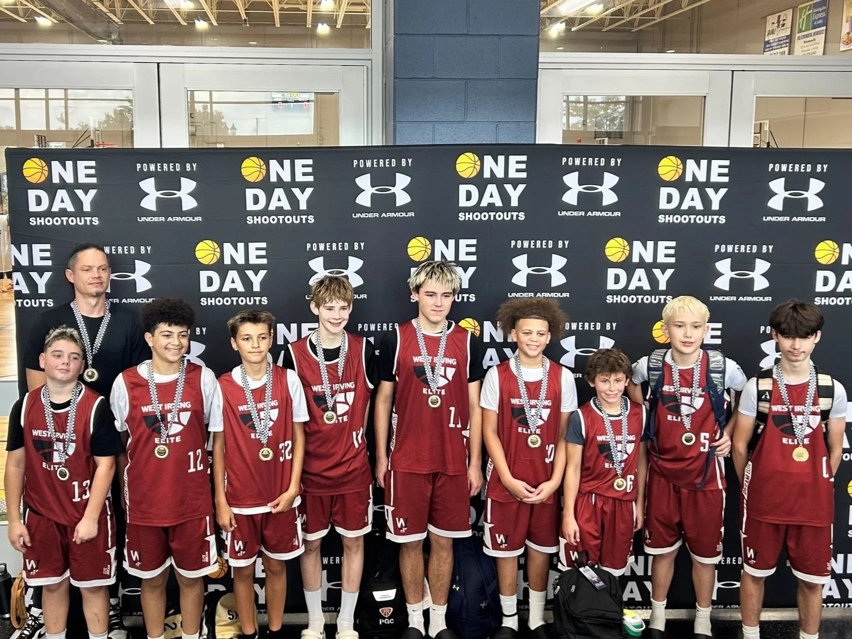 A group of young boys in red basketball uniforms and medals around their necks, standing in front of a black backdrop with logos and text, posing for a photo after a basketball tournament.