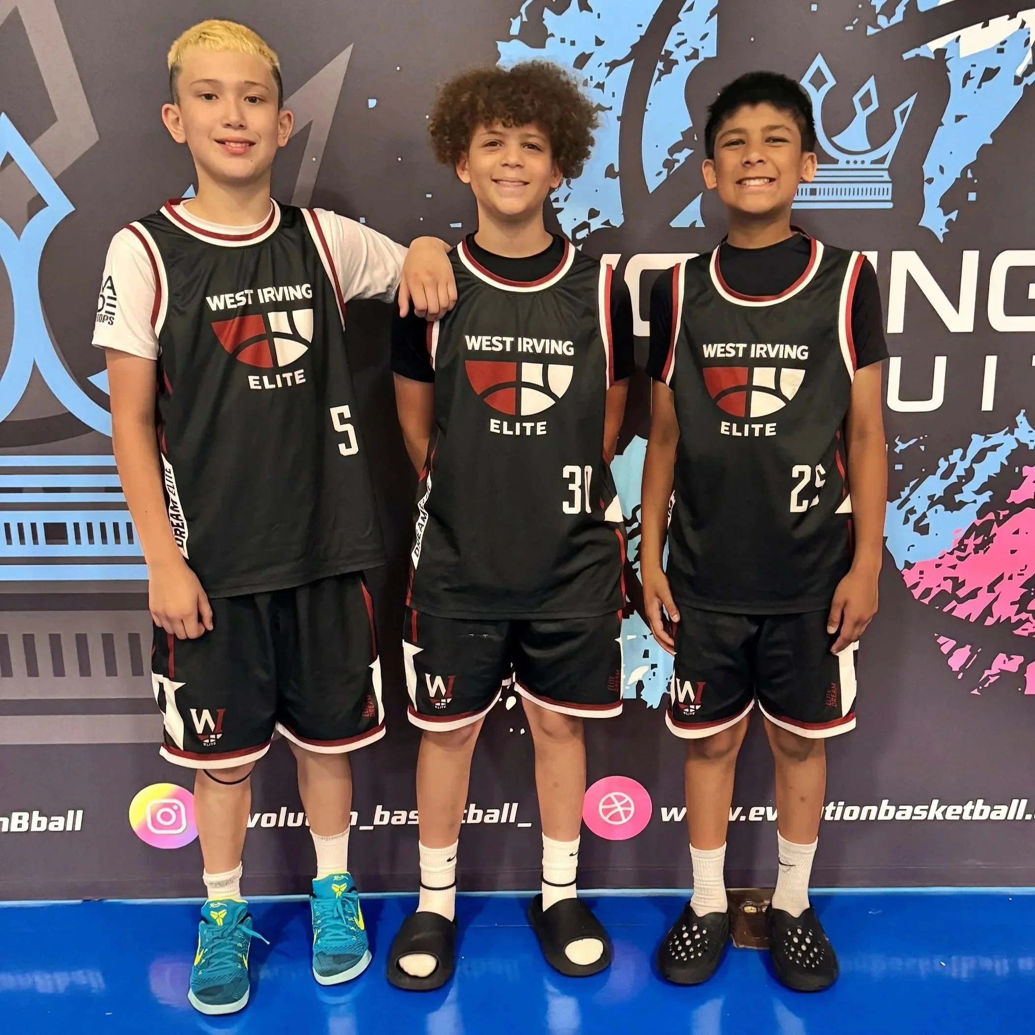 Three young boys standing side by side in basketball uniforms, smiling and posing for the camera against a colorful background.