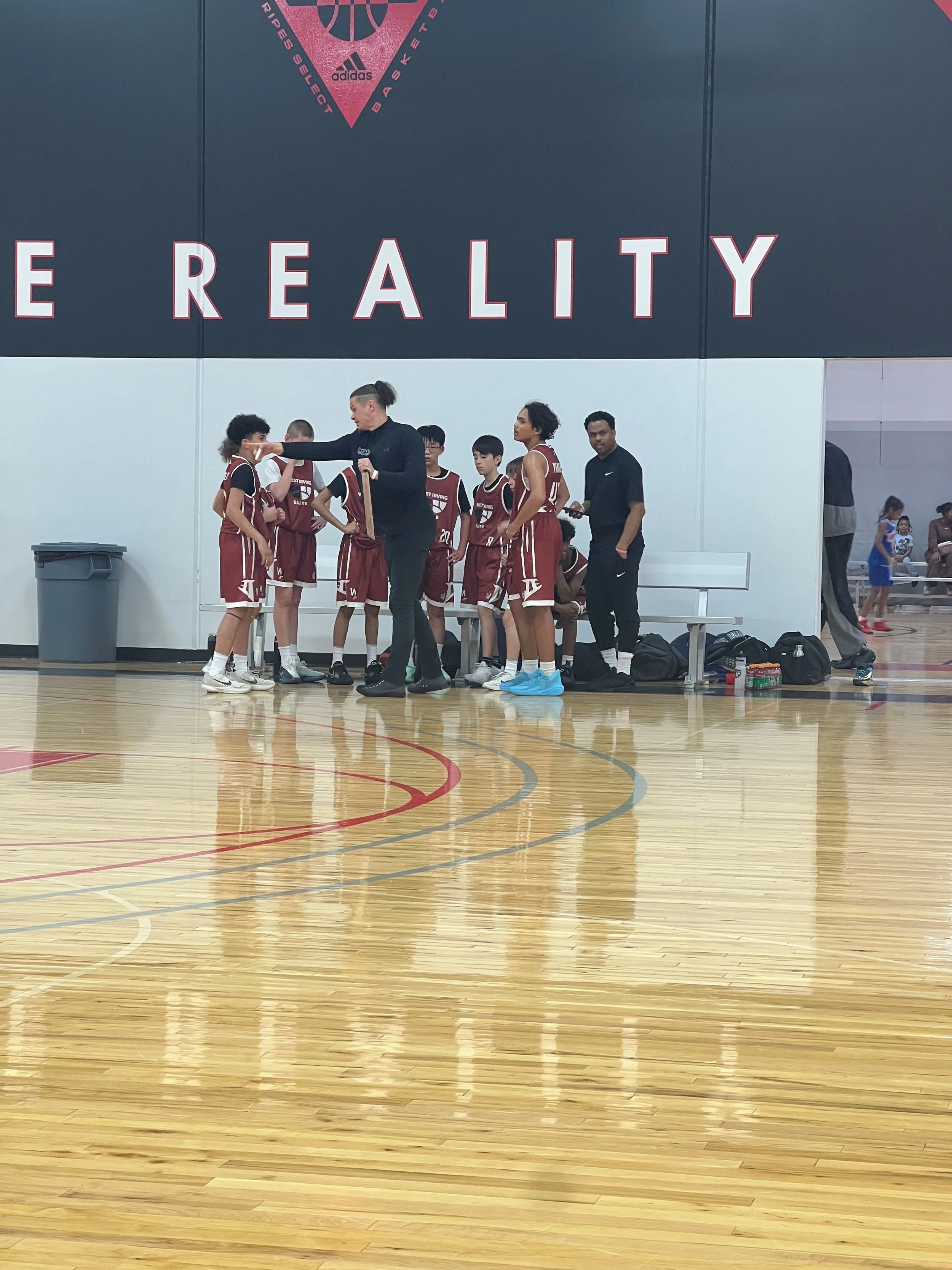 A basketball team coaching session taking place in an indoor gym. The coach, dressed in black, is talking to the players, who are in maroon jerseys and shorts, some with hands on hips, some listening attentively. There are bags and water bottles on the floor, and the gym has a polished wooden floor with red and gray lines. In the background, a few spectators and other players are visible. A large wall sign says 'THE REALITY.'