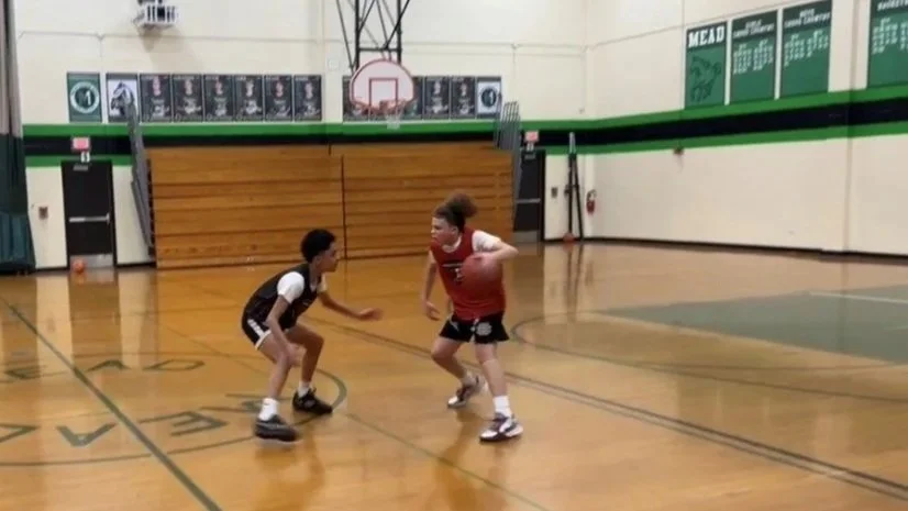 Two children playing basketball on an indoor court, with one dribbling the ball and the other in a defensive stance.