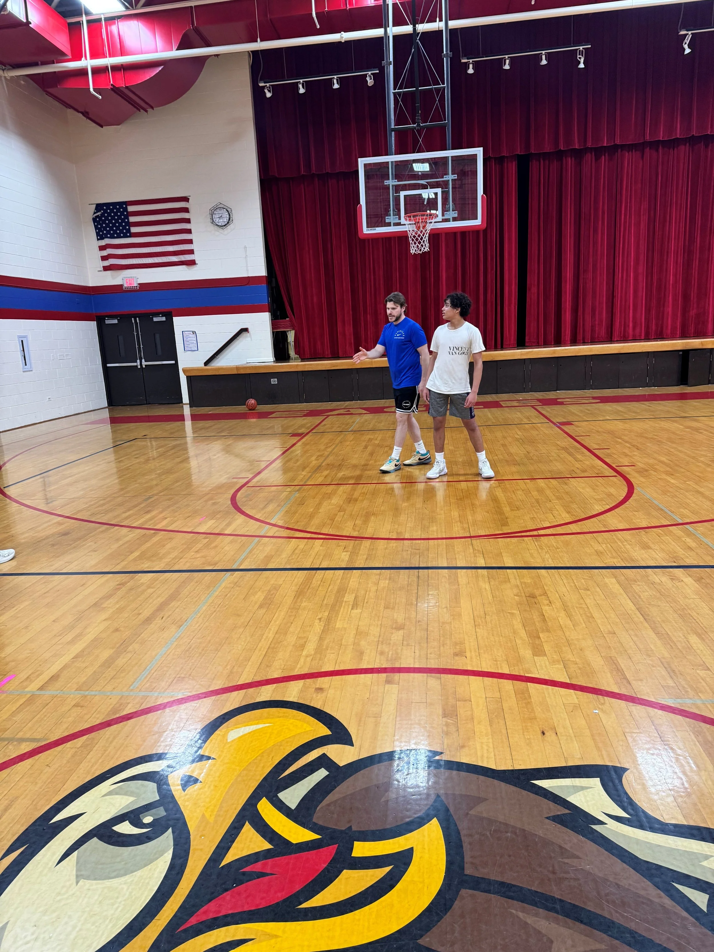 An indoor basketball court with a wooden floor, a basketball hoop, and two men standing and talking near the court. There is a logo on the floor and an American flag on the wall.