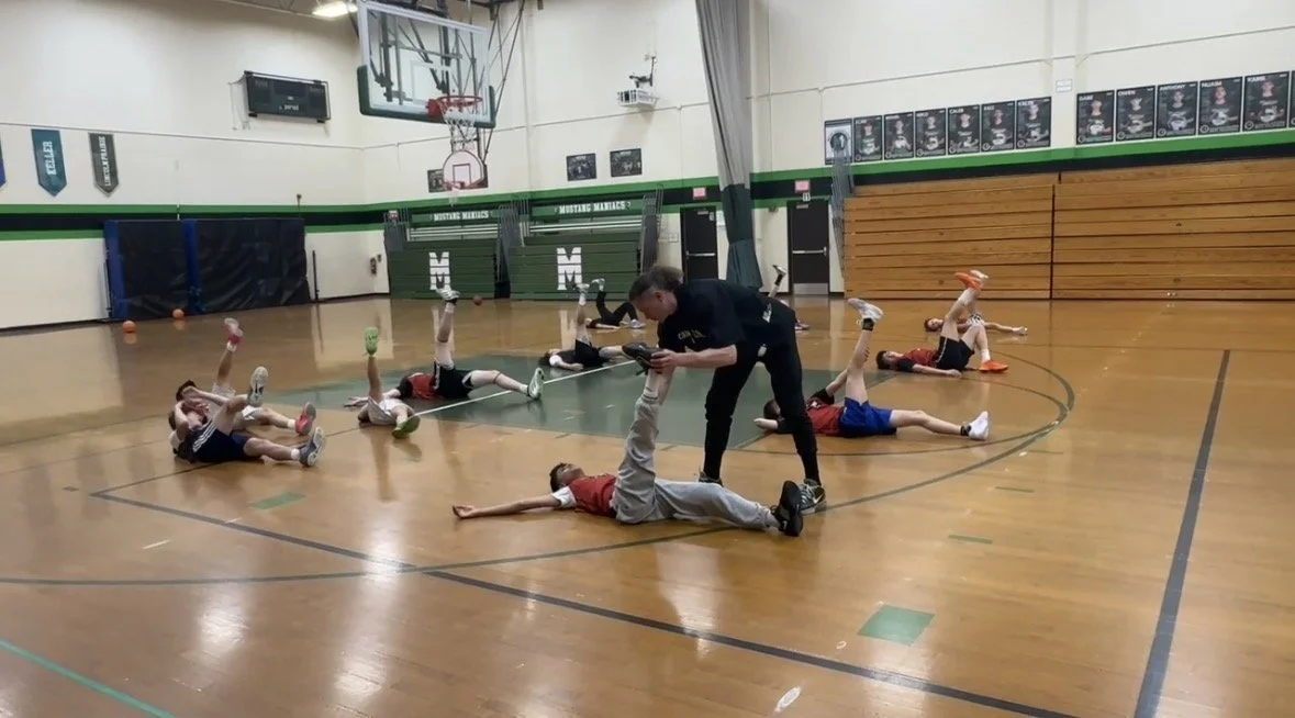 A group of young children lying on a basketball court, holding colorful balls in one hand with legs raised, practicing stretches or exercises, while a coach assists a child lying on the ground.
