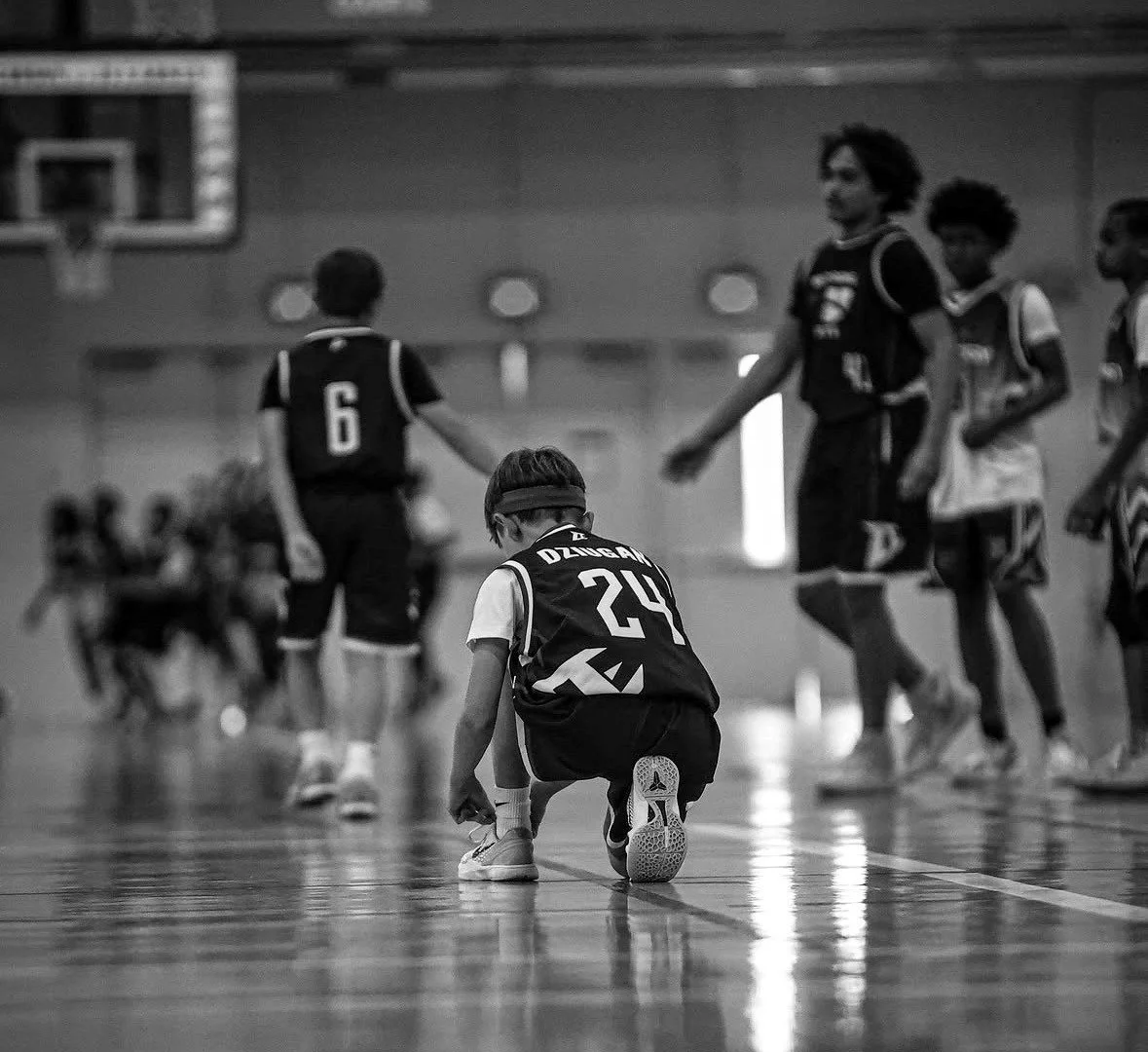 Young boy wearing a sports jersey and headband kneeling on the gym floor, with other children playing basketball in the background.