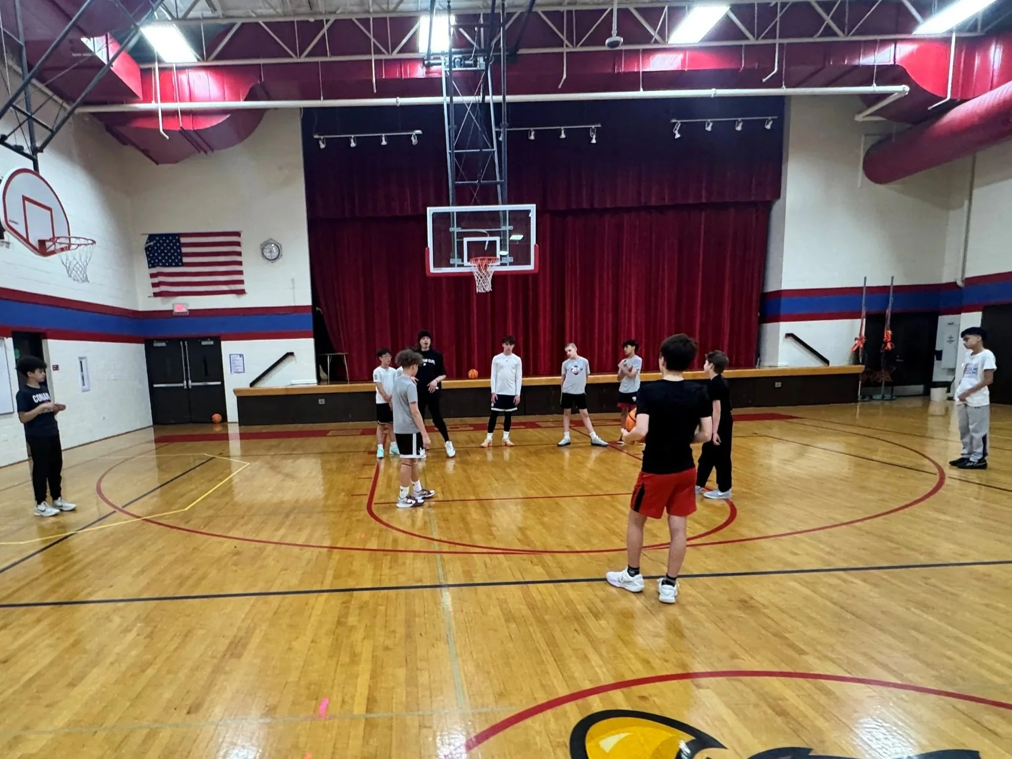 Students are gathered in a gymnasium for a basketball practice or game, with some people on the ground and others standing around. The gym has a polished wooden floor, basketball hoops, an American flag on the wall, and red curtains behind the hoop.