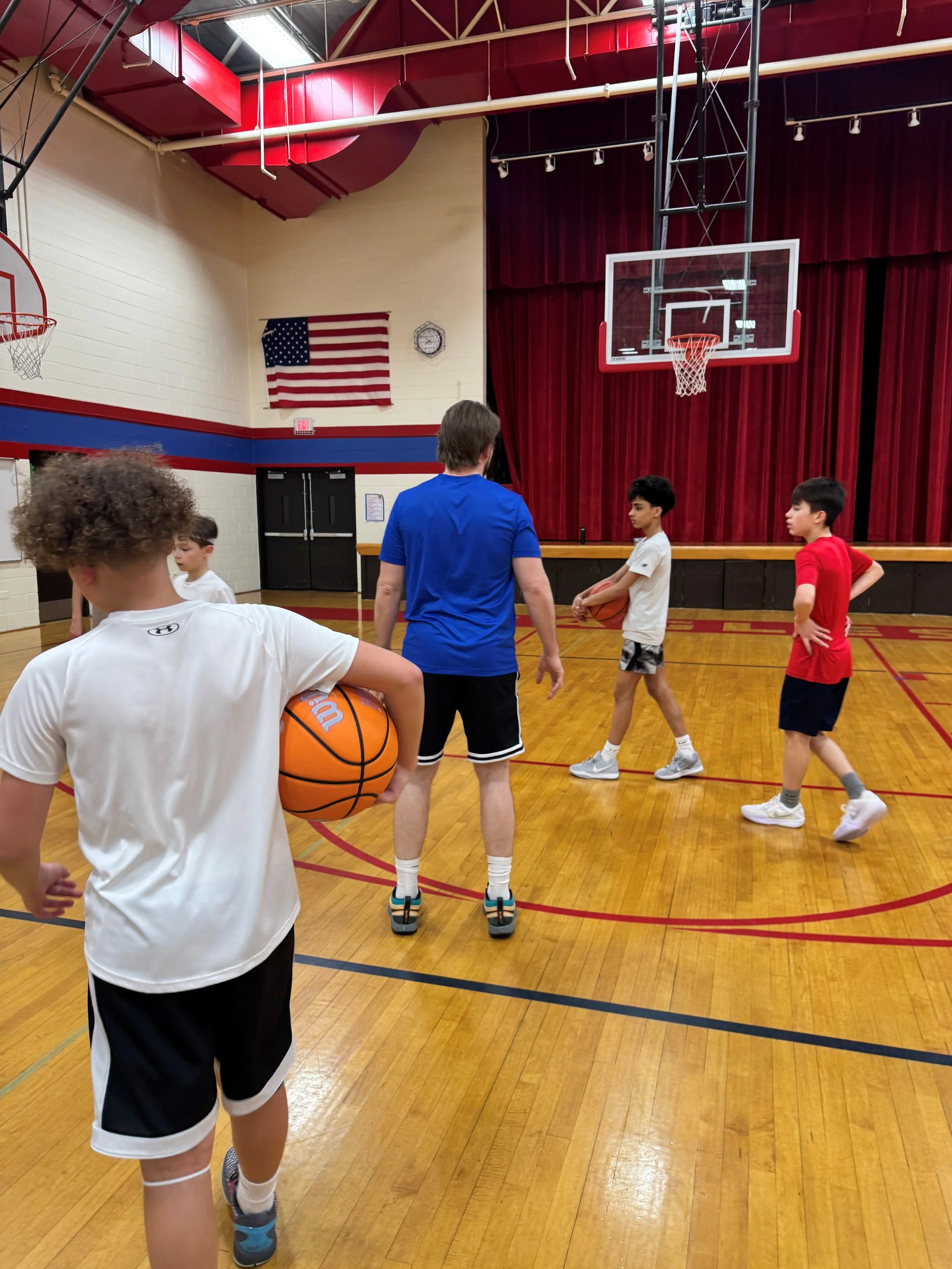 Children and a coach in a gymnasium. One child holds a basketball, others are standing and talking. The gym has red and blue walls, a basketball hoop, an American flag, and red curtains.