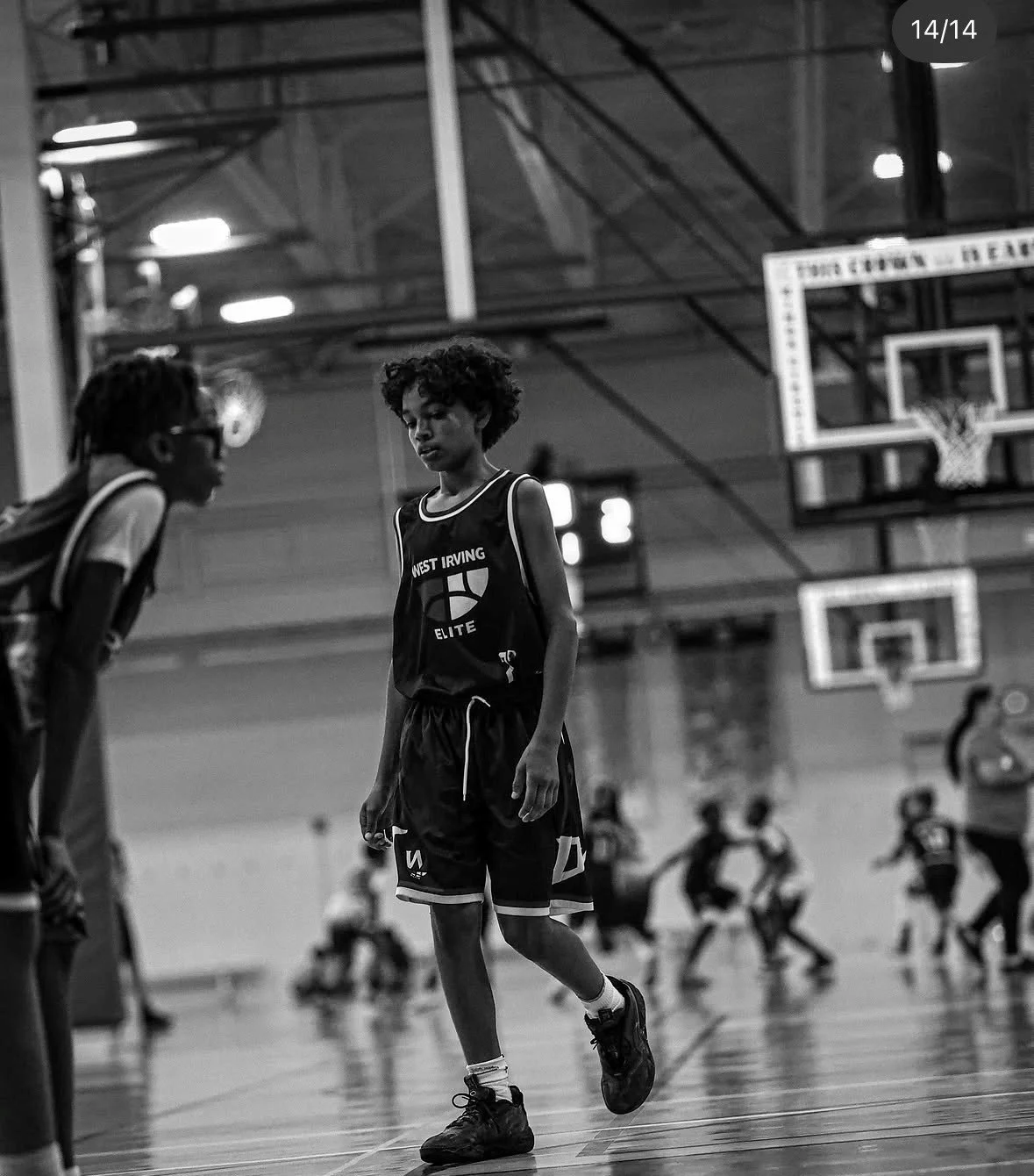 Black and white photo of a young basketball player standing on the court with other children playing in the background, inside a gymnasium with basketball hoops and overhead lights.