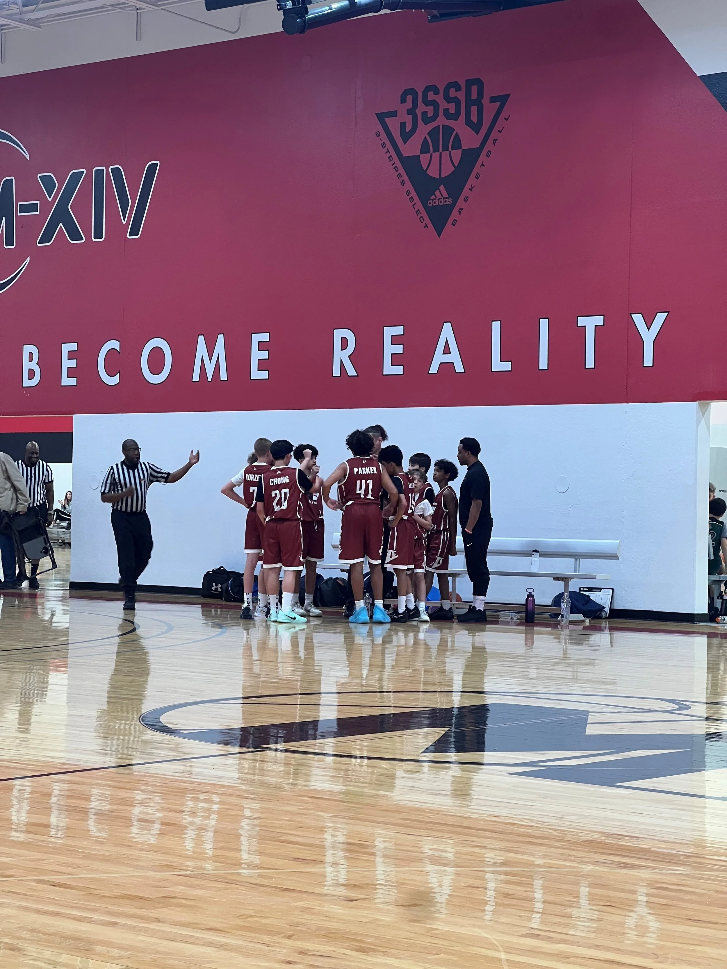 A group of young basketball players in maroon uniforms gathered around a coach or referee during a game timeout on a polished indoor basketball court. A large maroon wall in the background displays the text 'BECOME REALITY' and a Adidas team logo.
