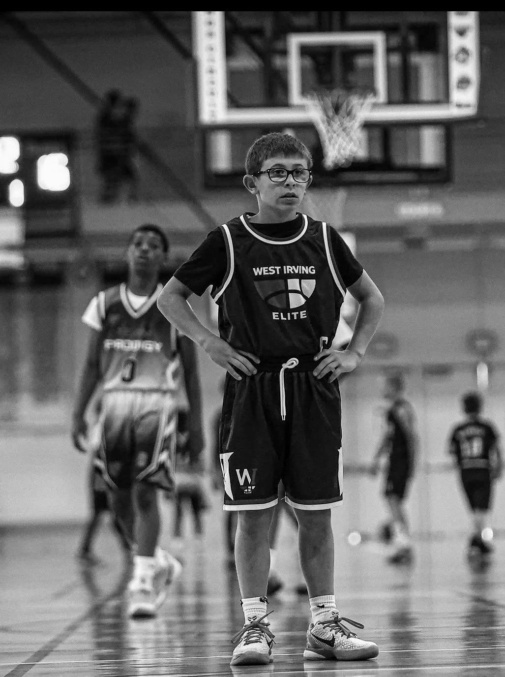 Young boy with glasses and short hair standing with hands on hips on a basketball court, wearing a 'West Irving Elite' jersey and shorts, with a basketball hoop in the background. Other children are present in the background.