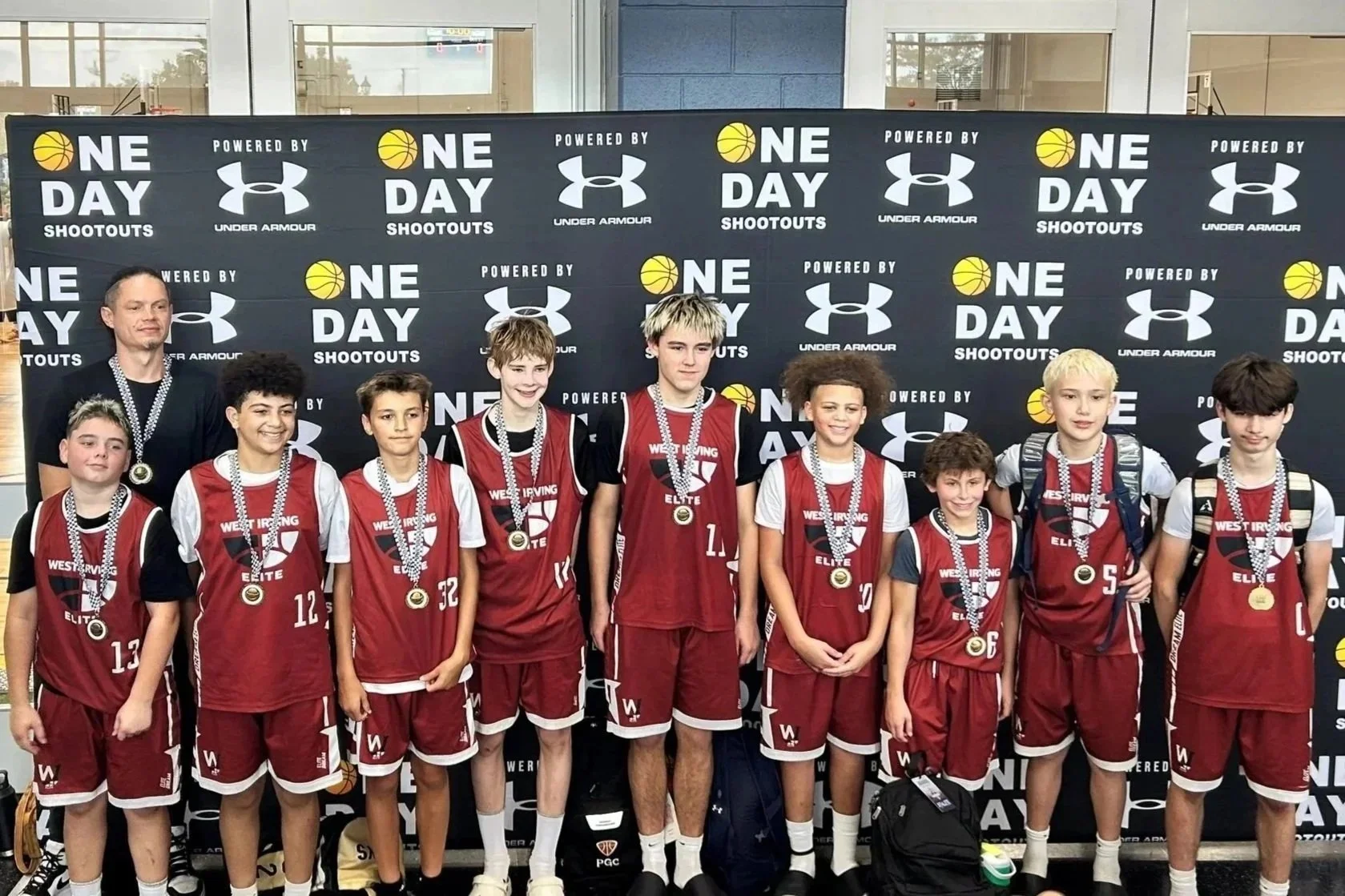 A youth basketball team wearing red uniforms and medals stands in front of a black backdrop with the logos 'One Day Shootouts' and 'Under Armour.' An adult coach or organizer stands to the left with a medal.