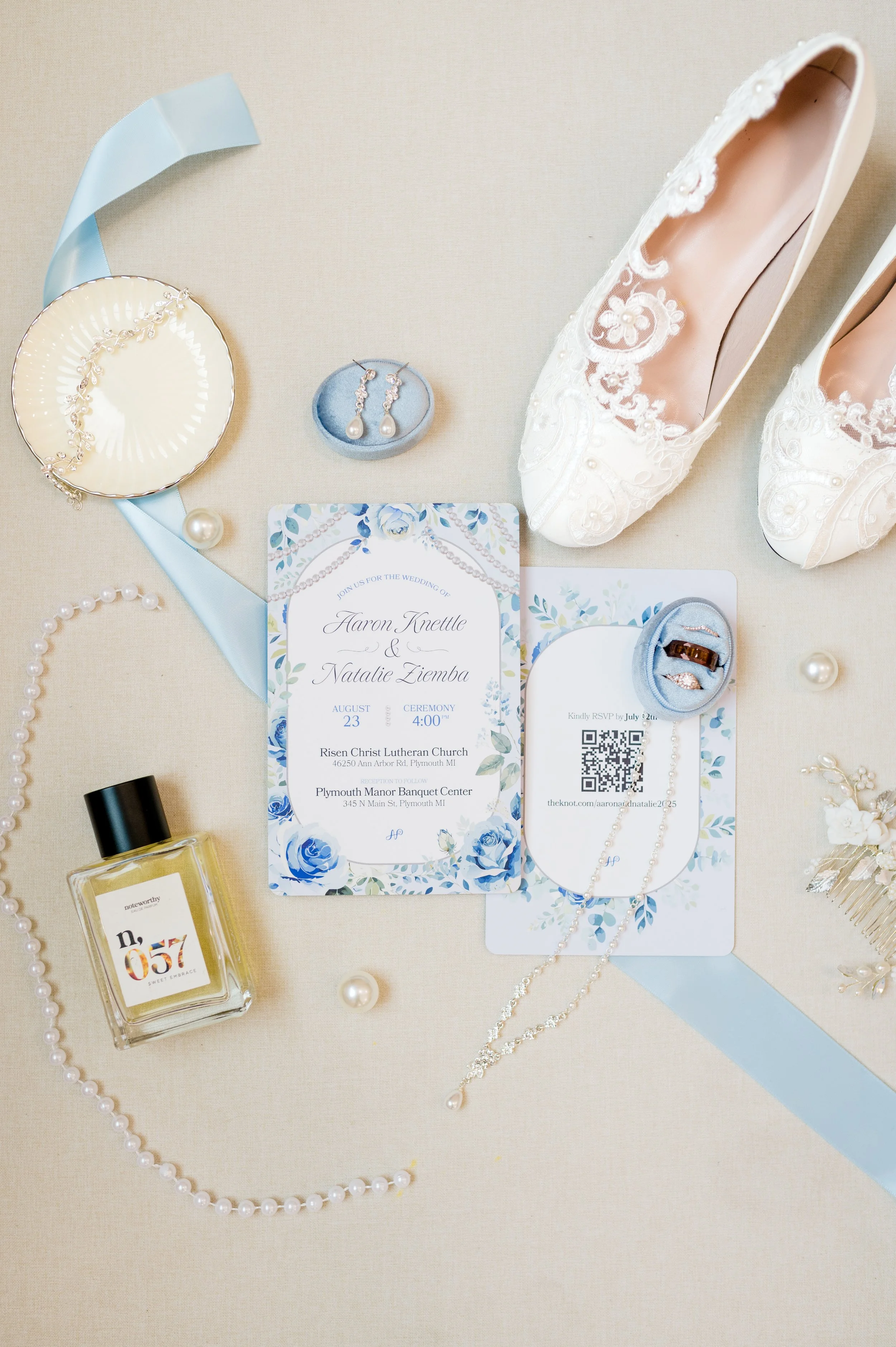 Wedding invitation surrounded by pearl necklace, pearl earrings, lace shoes, perfume bottle, and other jewelry on a light-colored surface.