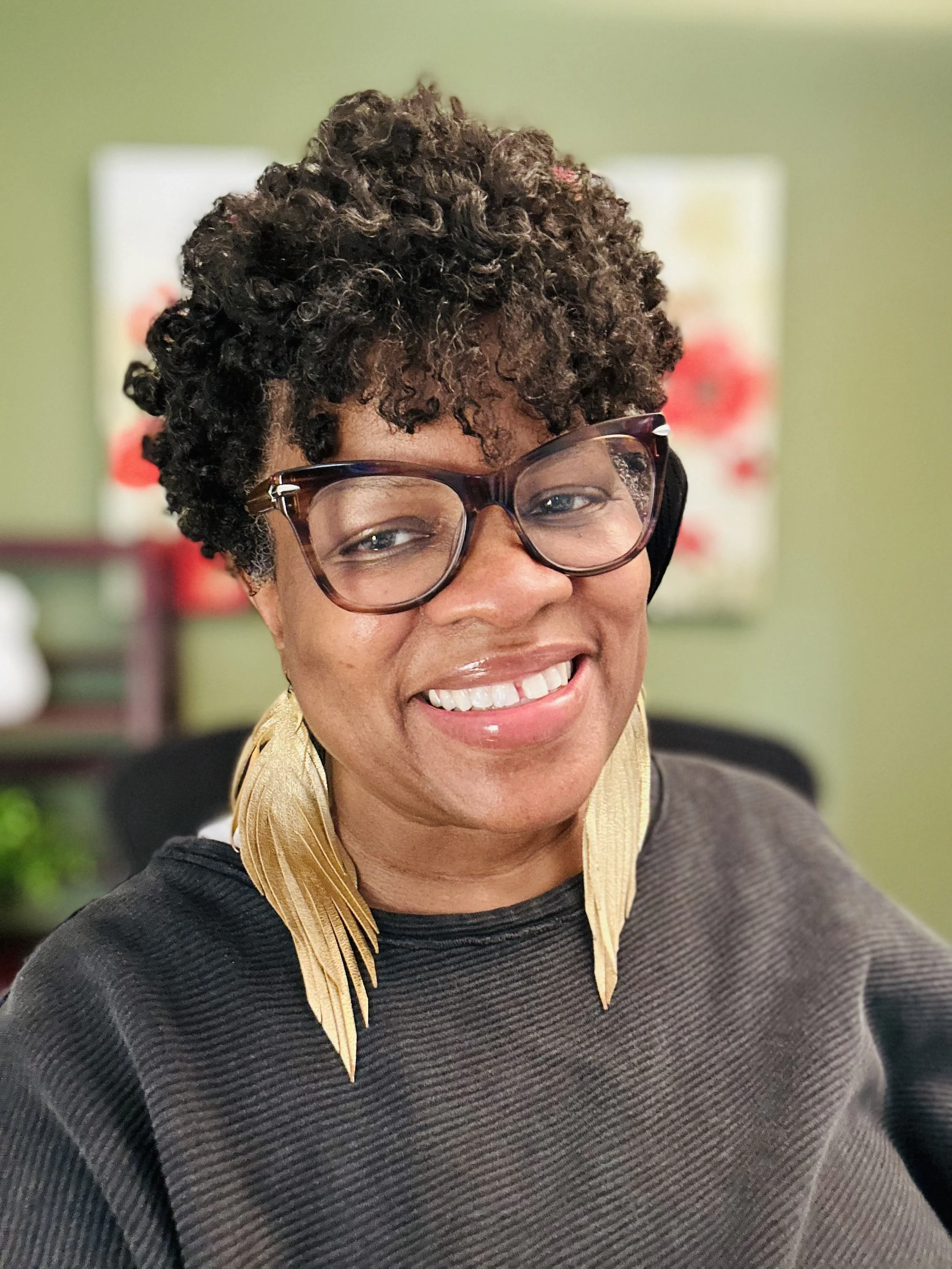 A smiling woman with short curly hair, wearing glasses, a black top, and a gold scarf, in an indoor setting with a green wall and artwork in the background.