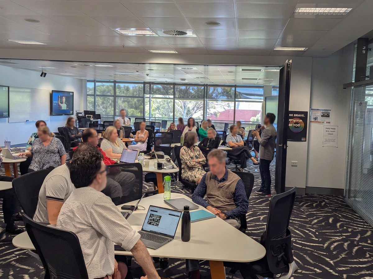A group of people attending a meeting or presentation in a conference room with large windows, some seated at tables with laptops, and a speaker standing near the entrance.