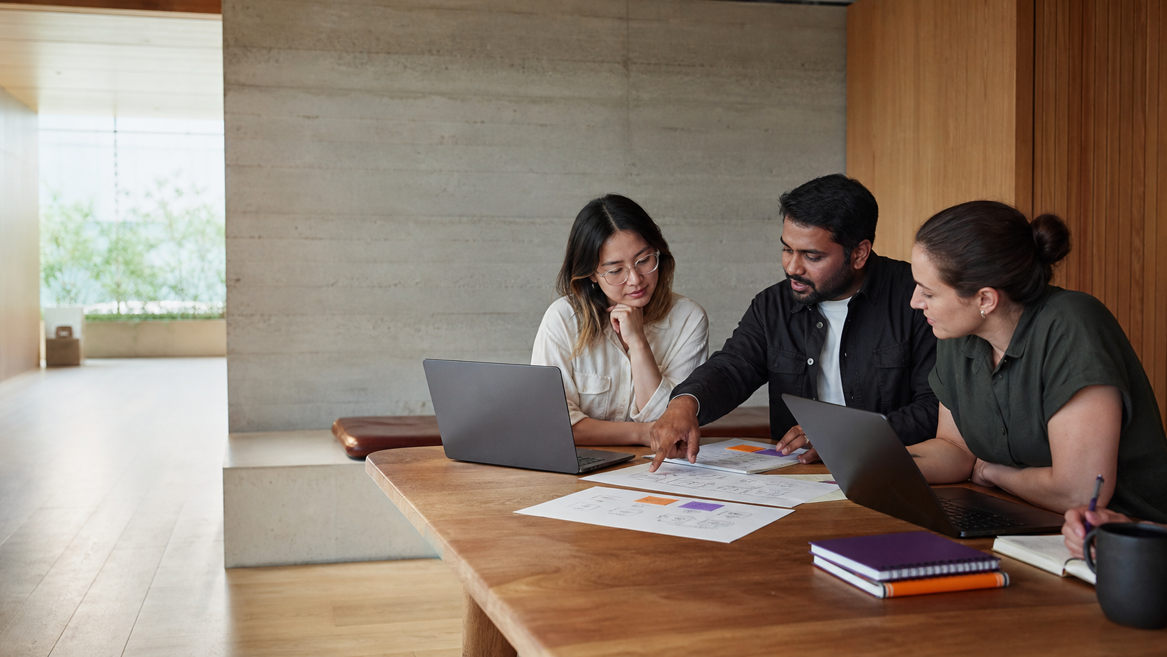 Three people sitting at a wooden table with laptops and documents, engaged in a discussion, in a modern office space with natural light.