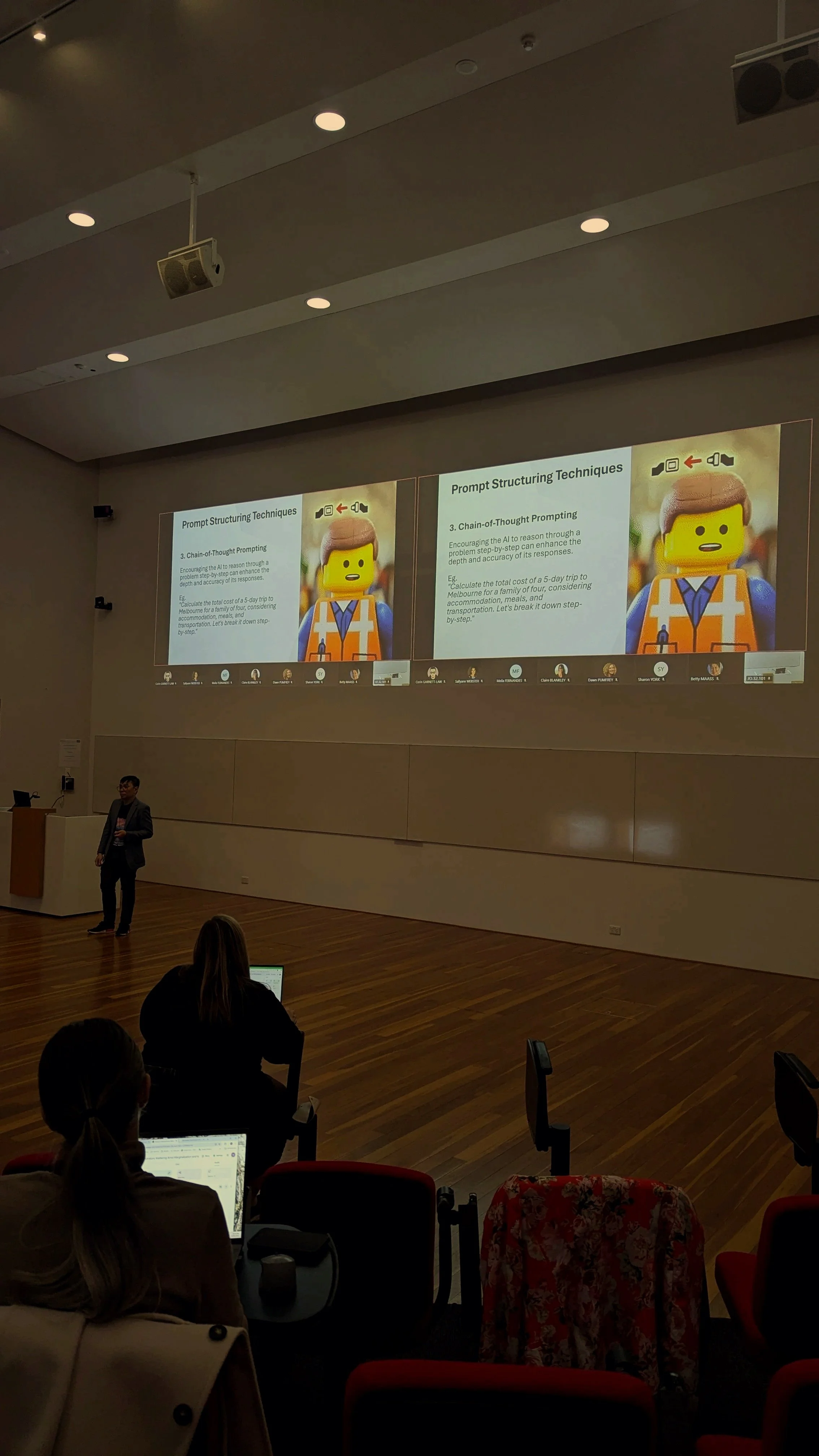 Lecture hall with people seated, presenter standing by a podium, large screen displaying a slide titled 'Prompt Structuring Techniques' with a subheading '3. Chain-of-Thought Prompting' and an image of a LEGO figure dressed as a construction worker.