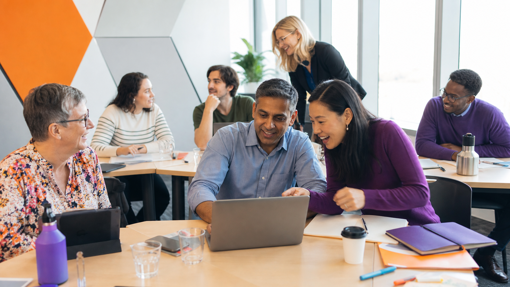 A group of seven diverse people in a modern office, engaging and smiling while looking at a laptop, with others in the background around a table, during a meeting or collaborative work session.