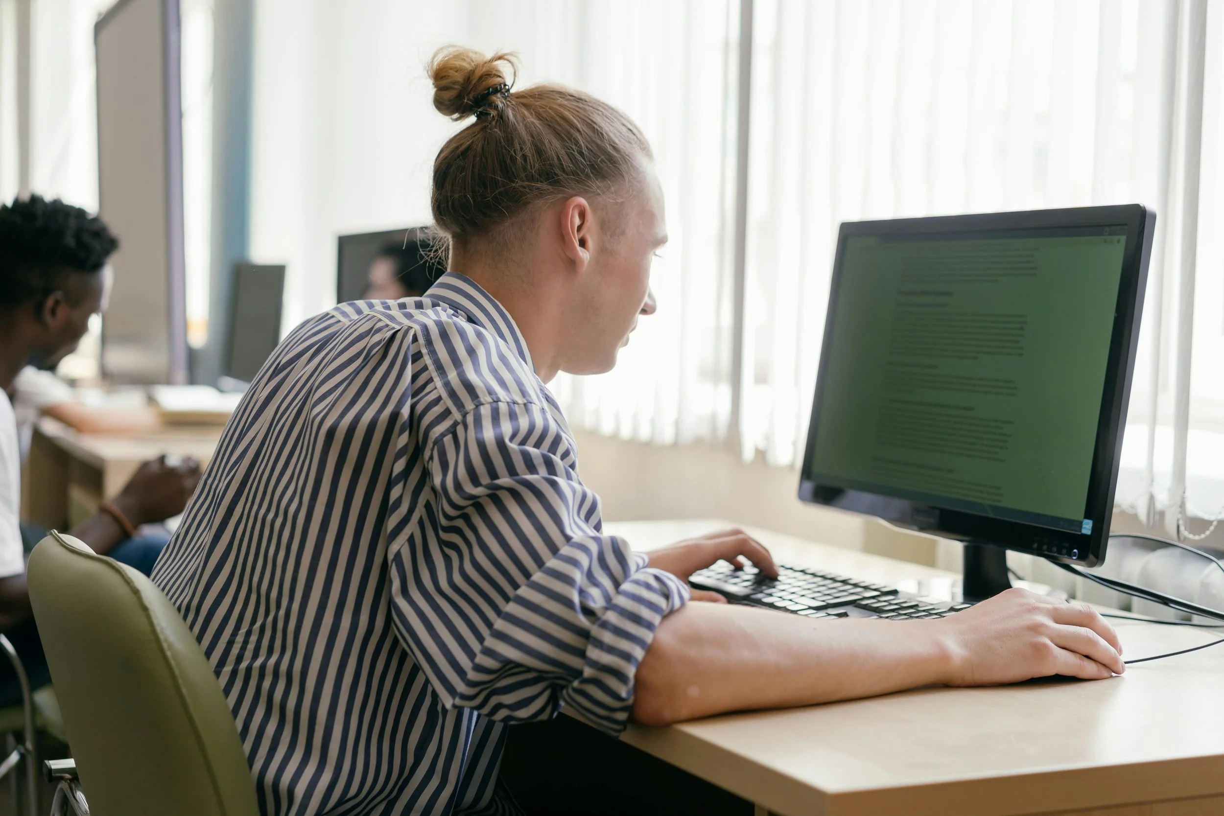 A young man with a top bun hairstyle sitting at a desk working on a computer in a well-lit room with large windows.