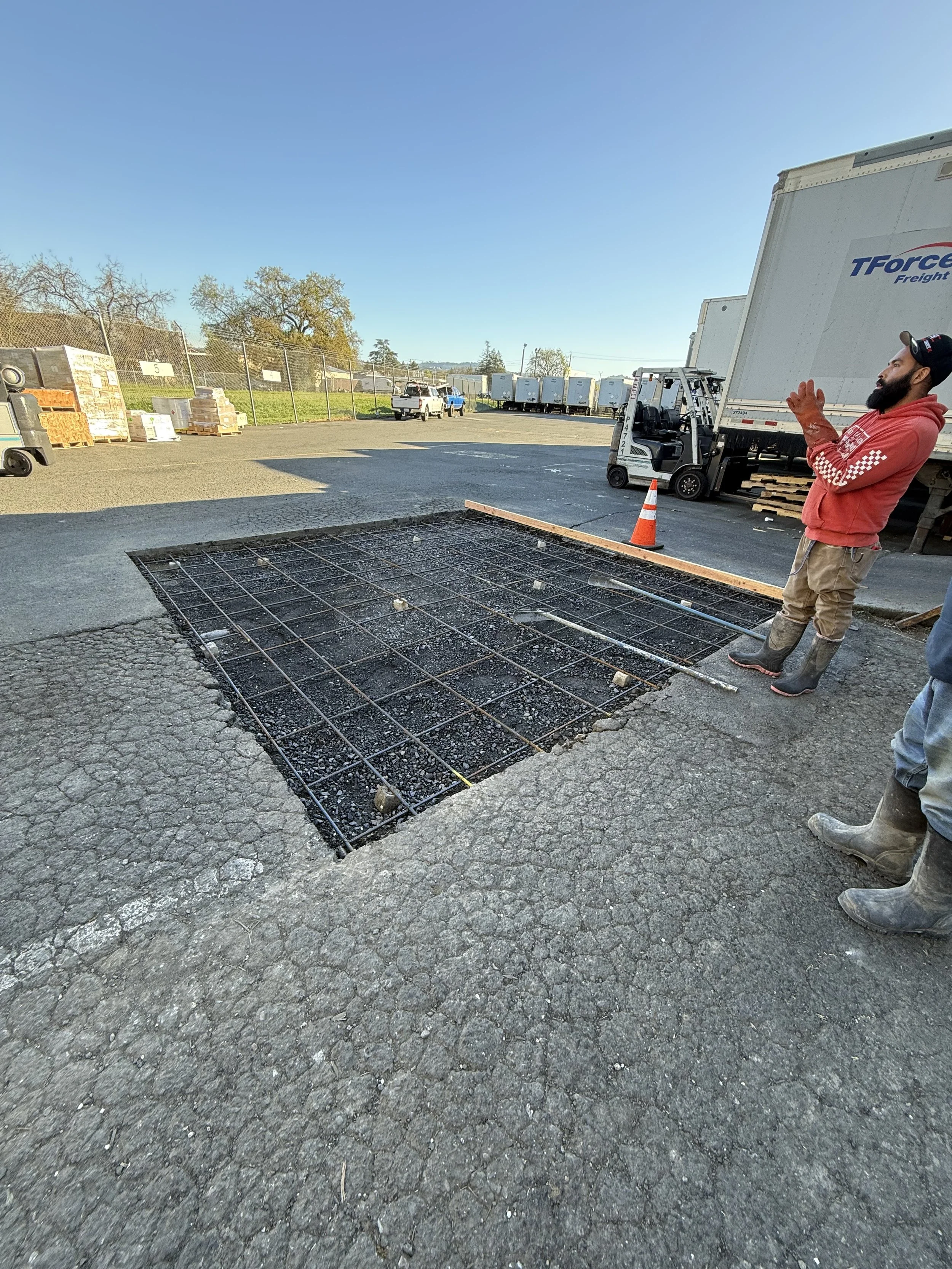 A construction worker standing next to a hole in the pavement with rebar reinforcement, orange safety cone, forklift, trucks, and parked cars in a lot under a clear blue sky.