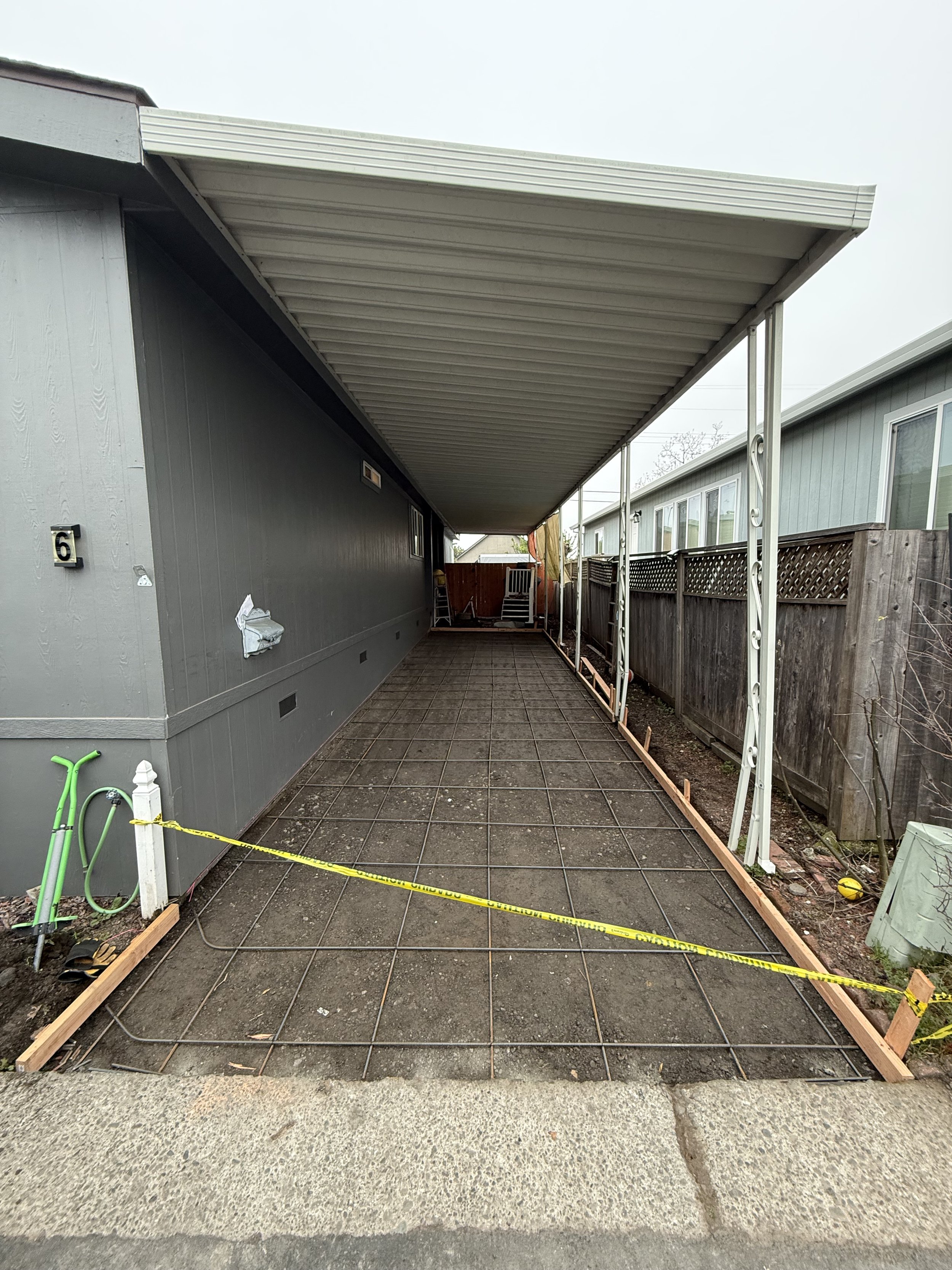 Back patio area with construction materials and equipment, including wooden framing, yellow caution tape, green garden hoses, and metal supports for a covered patio.