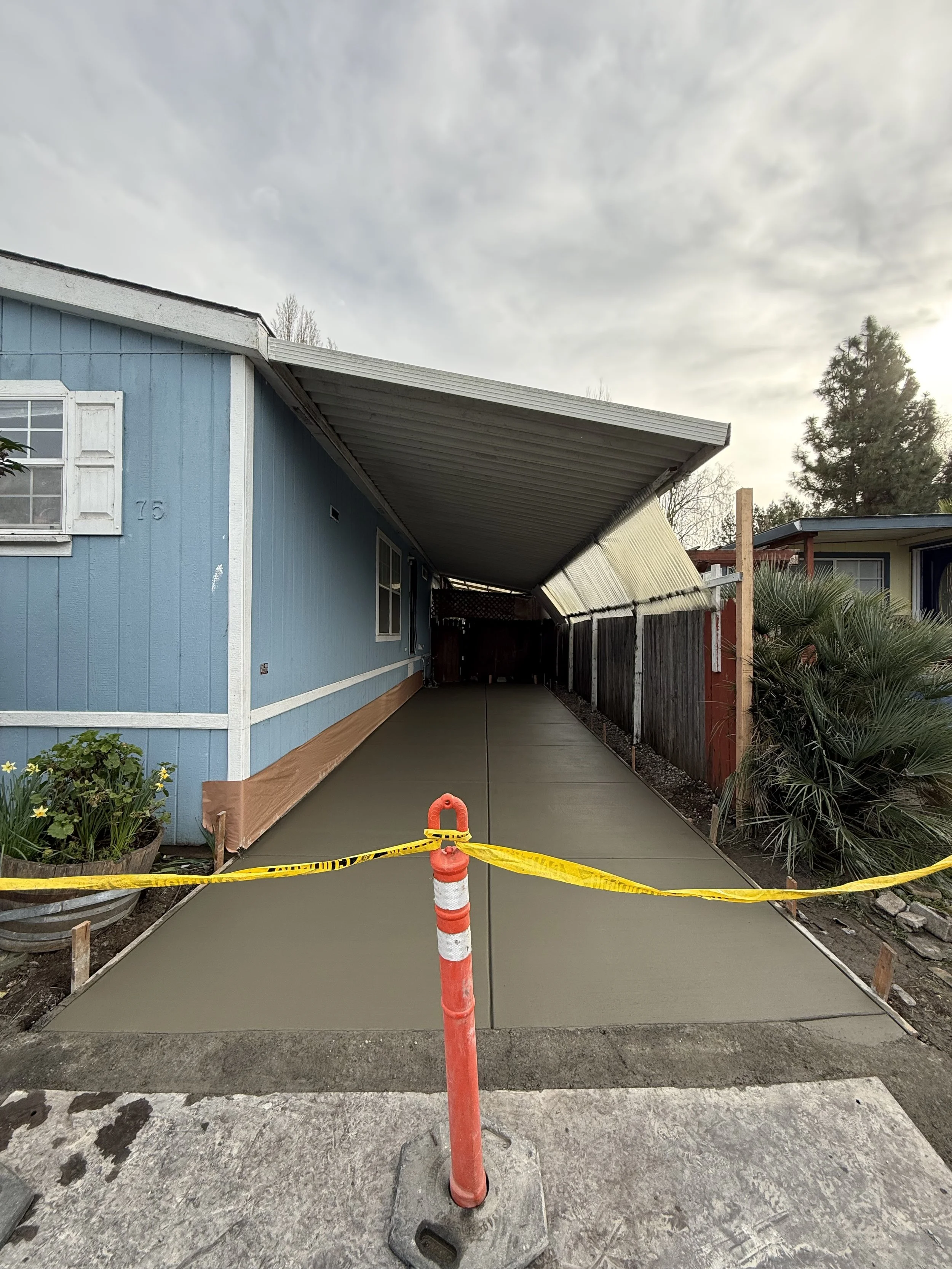 Newly poured concrete sidewalk section enclosed with yellow caution tape and orange traffic cone in front, adjacent to a blue house with white trim and window shutters, and an overhang roof partially covering the sidewalk.