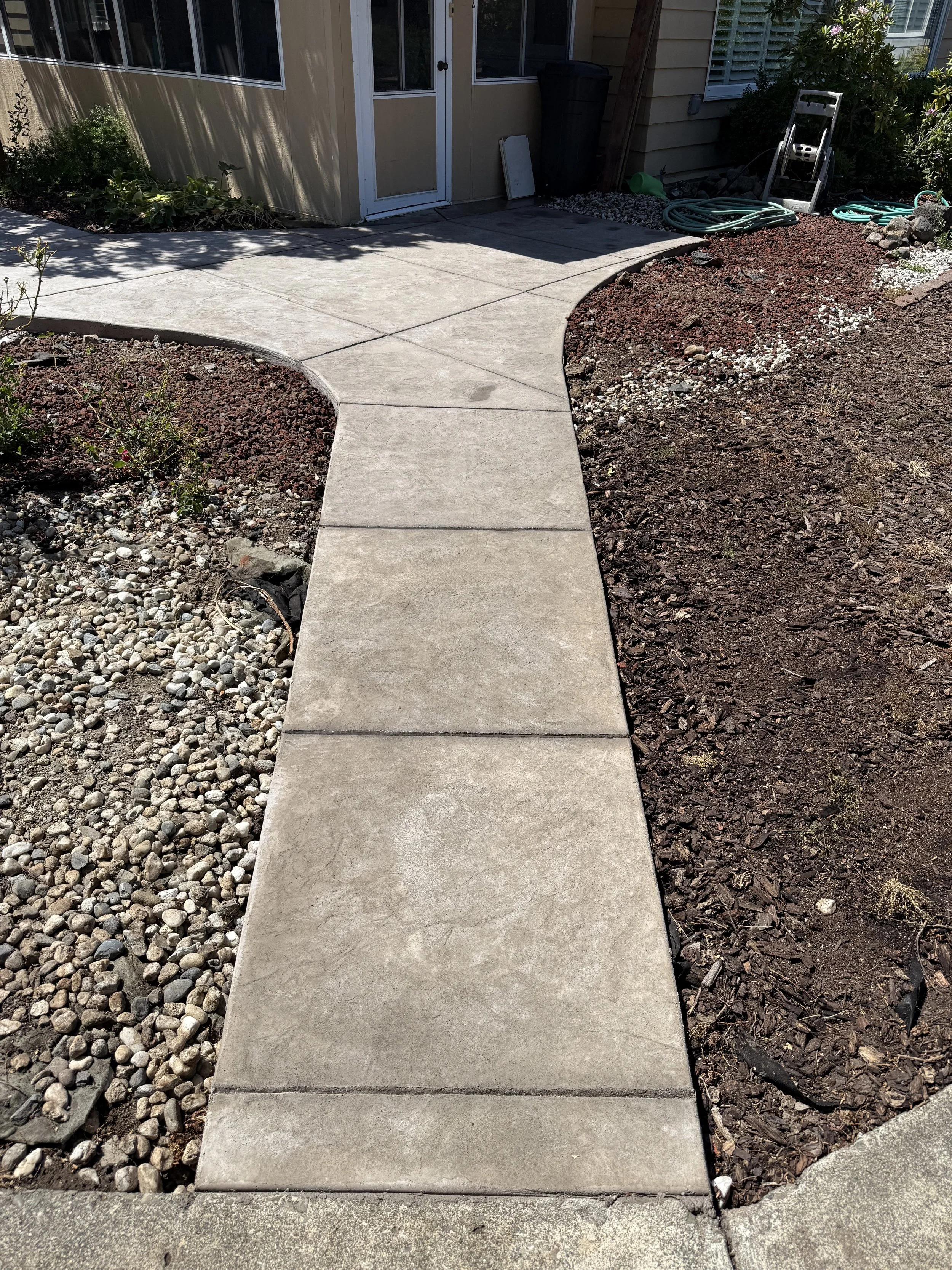 Newly installed concrete stepping stones forming a curved pathway leading to a door in a house. The surrounding yard has decorative rocks and mulch, with some gardening tools and hoses nearby.