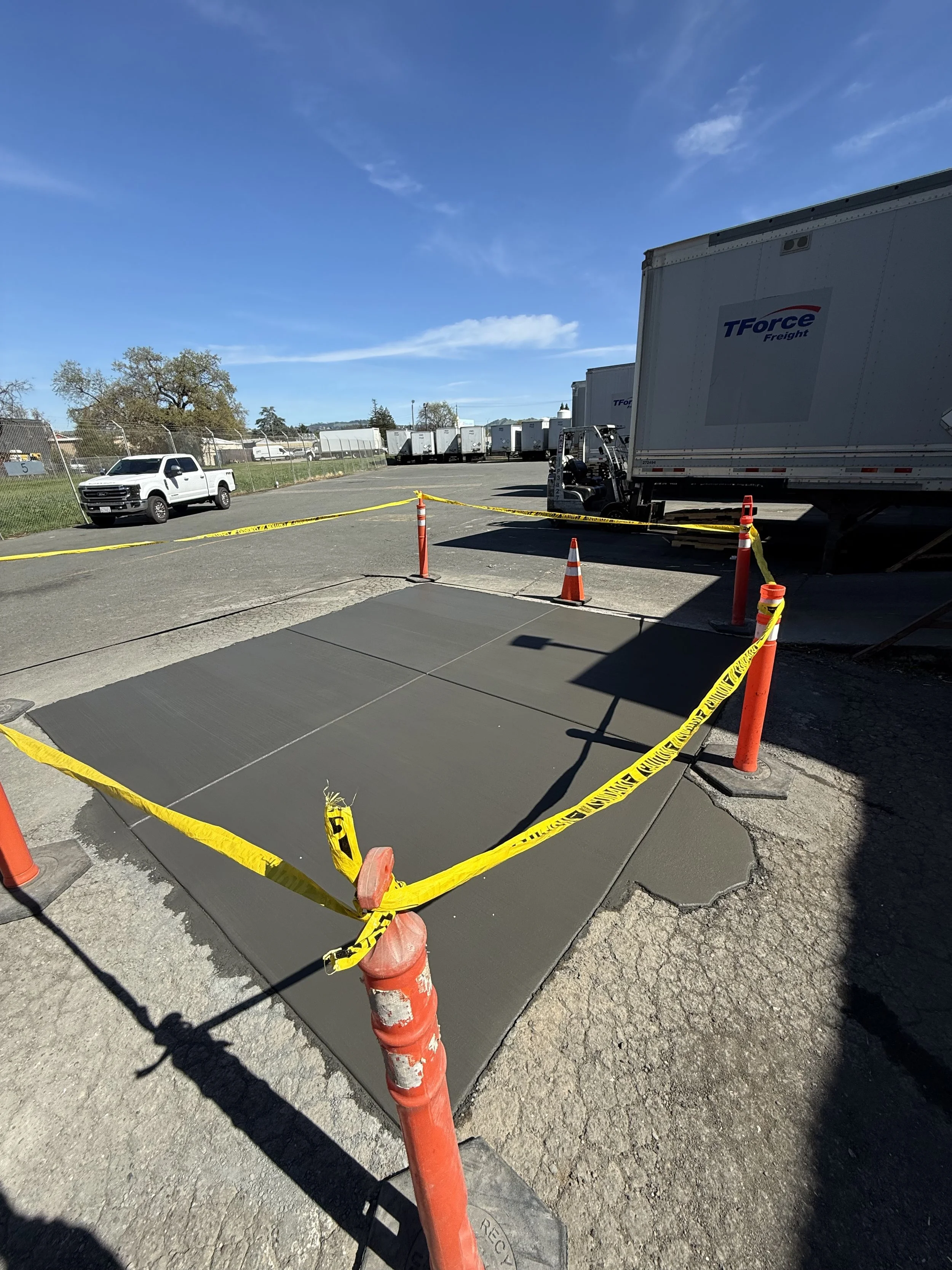Concrete slab being poured at a construction site surrounded by orange cones and yellow caution tape, with trucks and a forklift in the background under a clear blue sky.