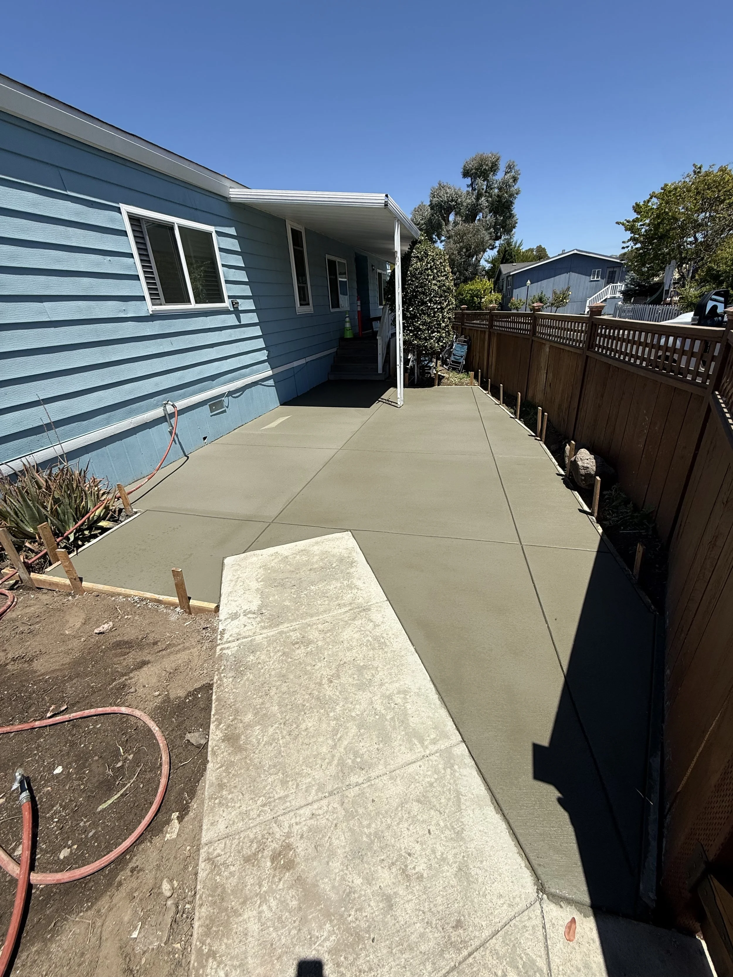 A newly poured concrete patio with smooth surface next to a house with blue siding, windows, and a small staircase, enclosed by a wooden fence, under a clear blue sky.