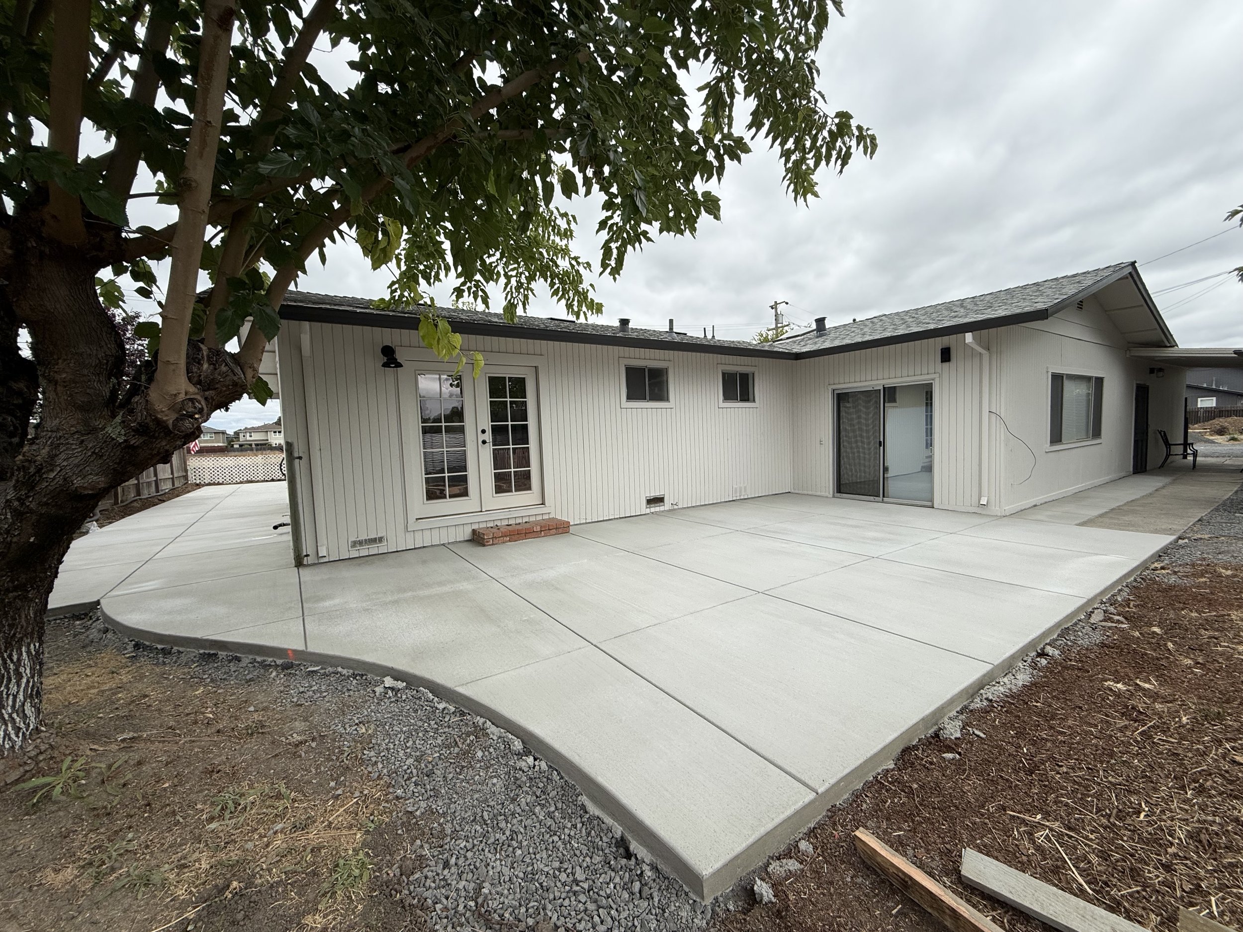 Backyard patio with new concrete slab and a tree in the foreground, house with sliding glass door and windows, overcast sky.