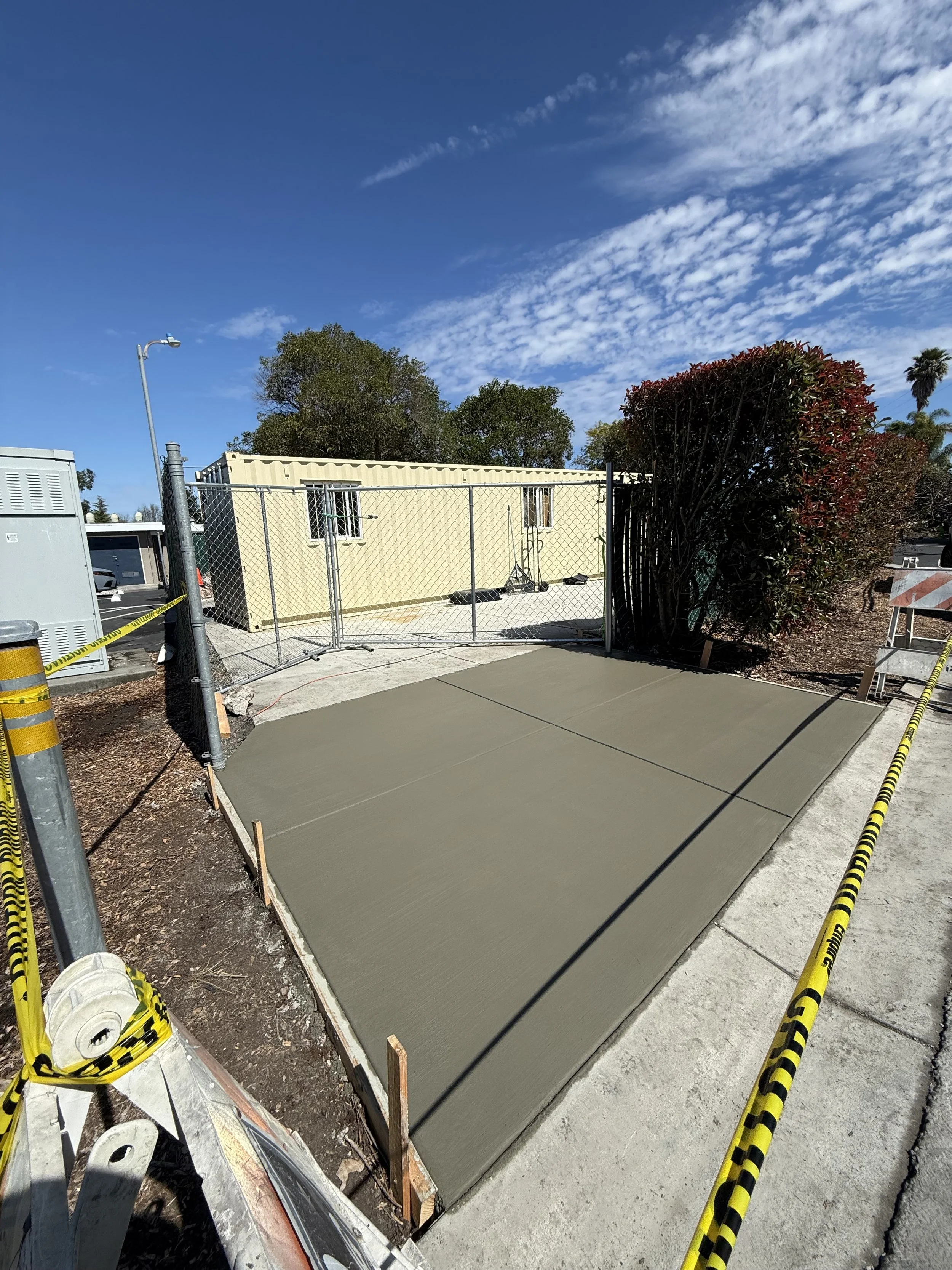 Freshly poured concrete on a sidewalk with construction caution tape and equipment nearby, fenced off for curing.