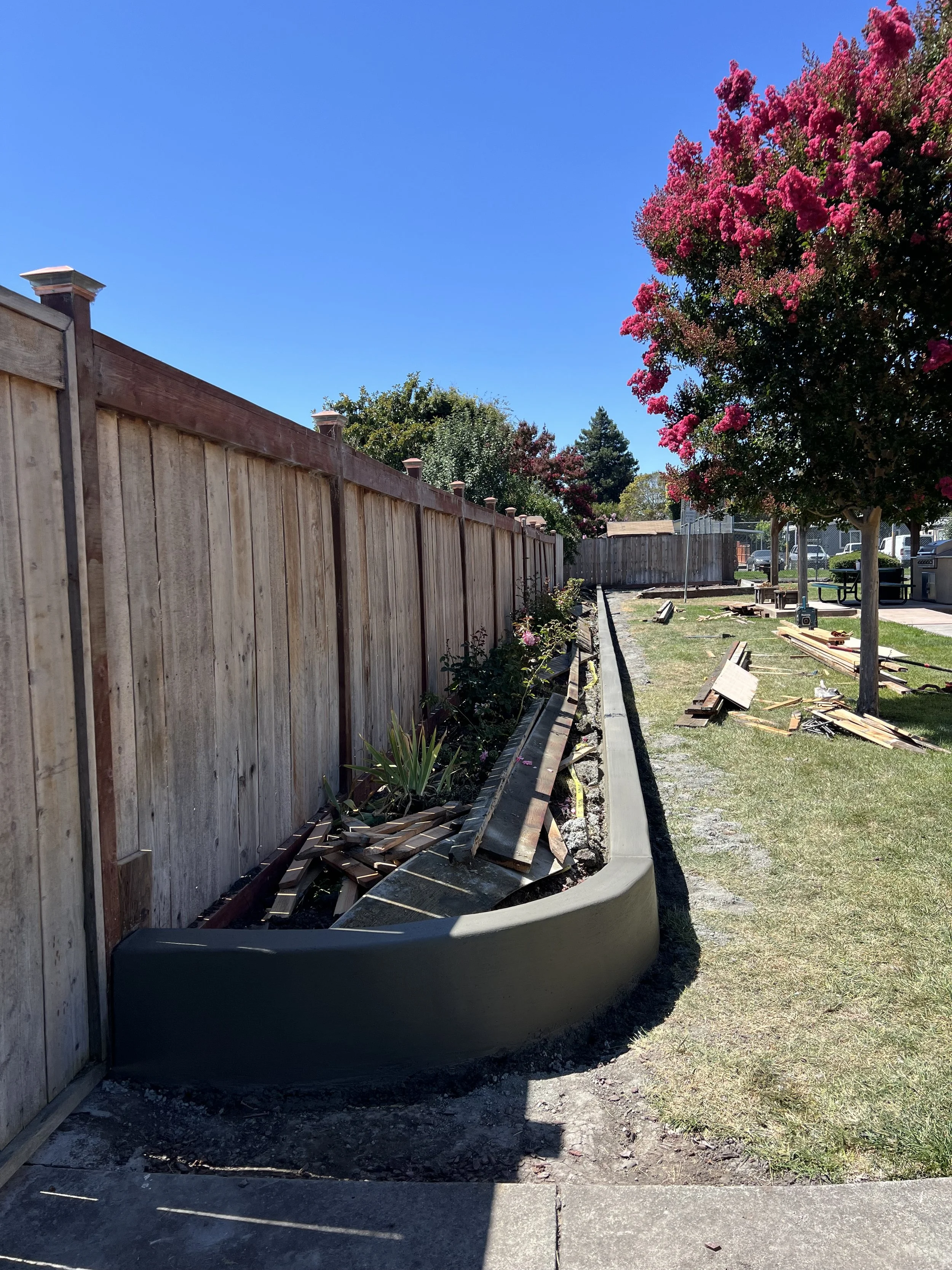 A wooden fence runs along the left side of a sidewalk, with some pink flowering trees on the right. Construction or landscaping work is ongoing, with wooden planks and construction materials scattered on the ground. The sky is clear and blue.
