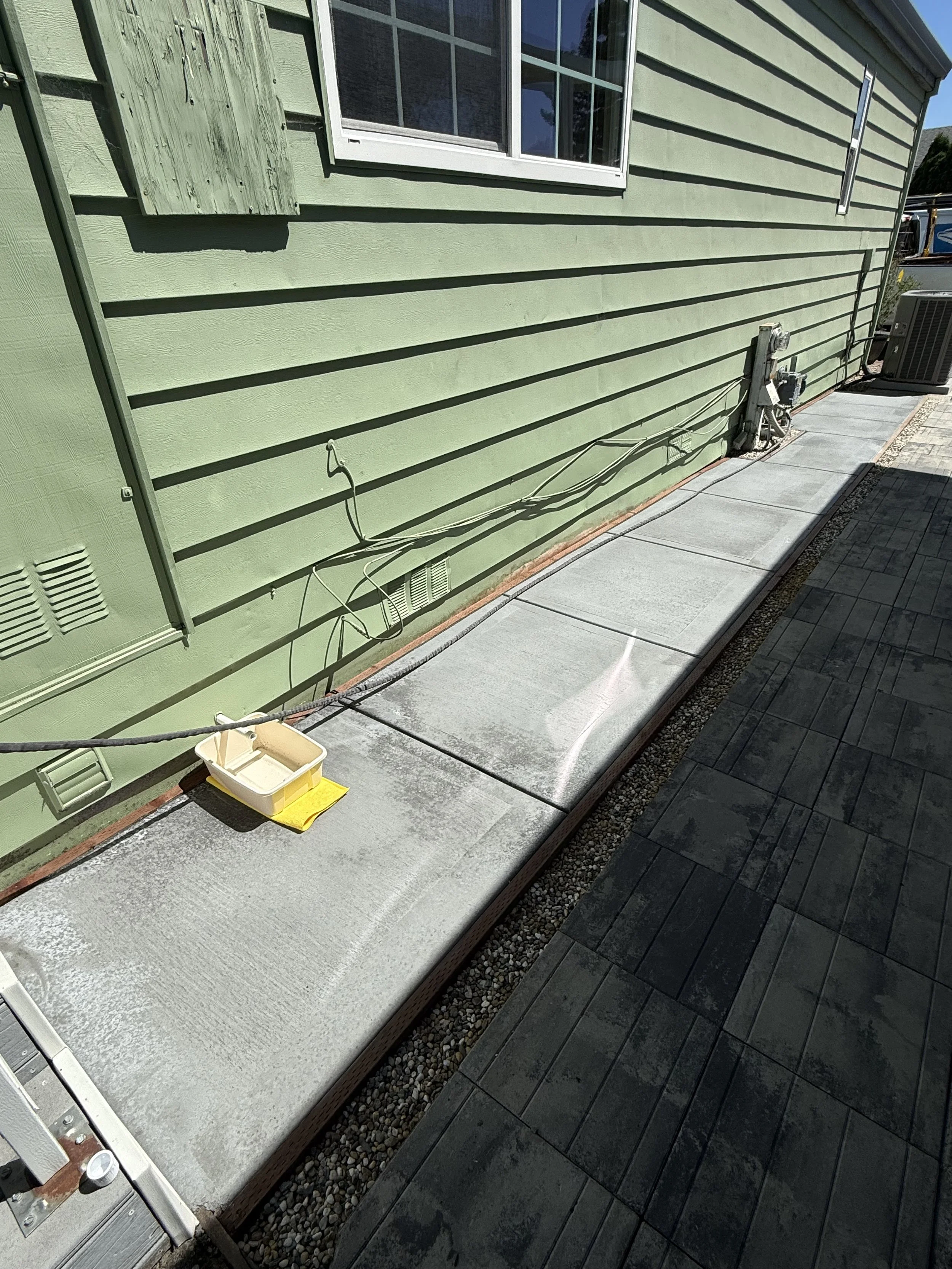 A side of a house with green horizontal siding, a white-framed window, and a shutter. There is a concrete walkway along the house with electrical outlets and visible wiring. A small bucket with a yellow cloth is placed on the walkway.