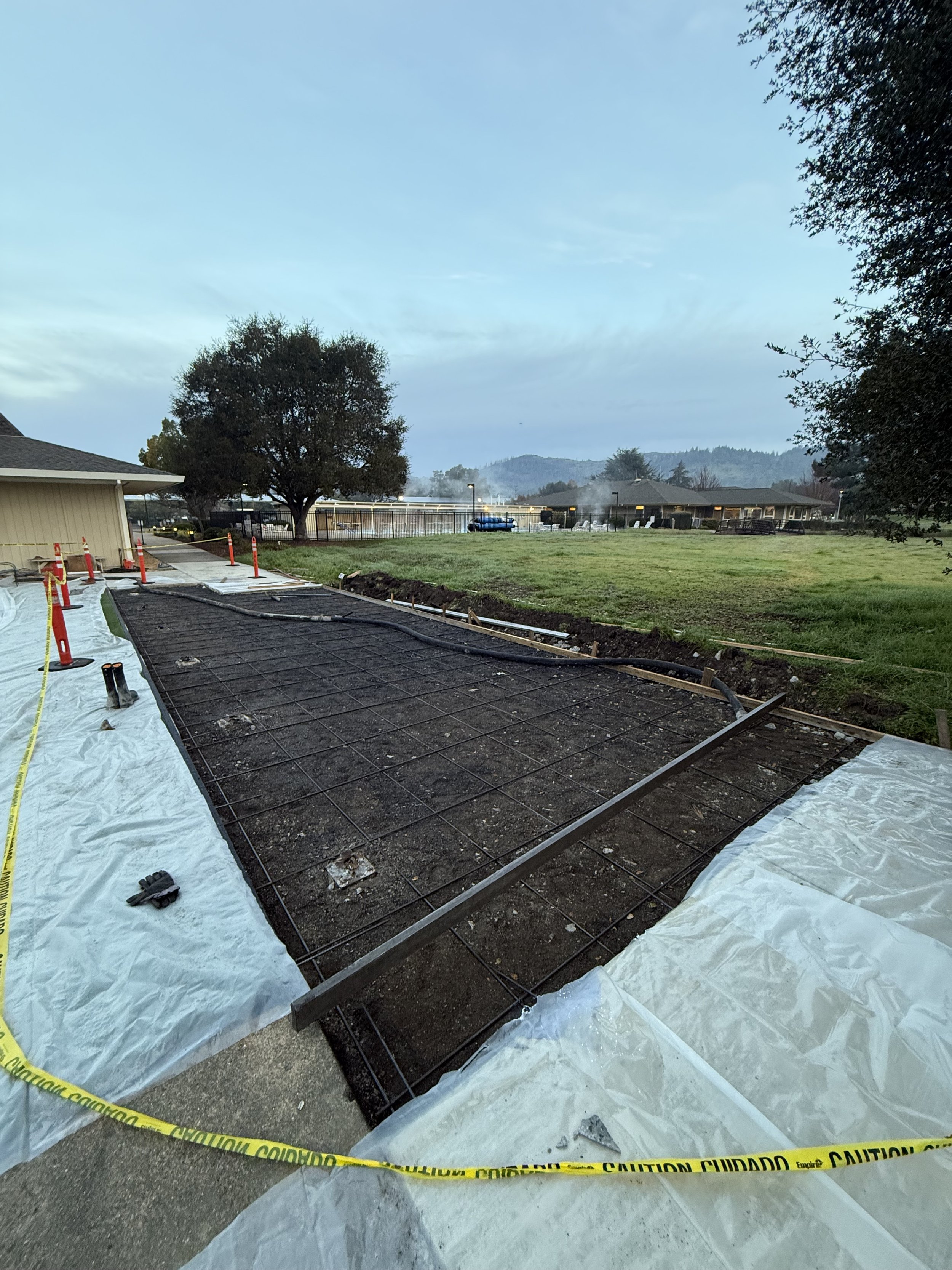 Construction site with exposed rebar, plastic sheeting, and caution tape, with a grassy area, trees, and buildings in the background.