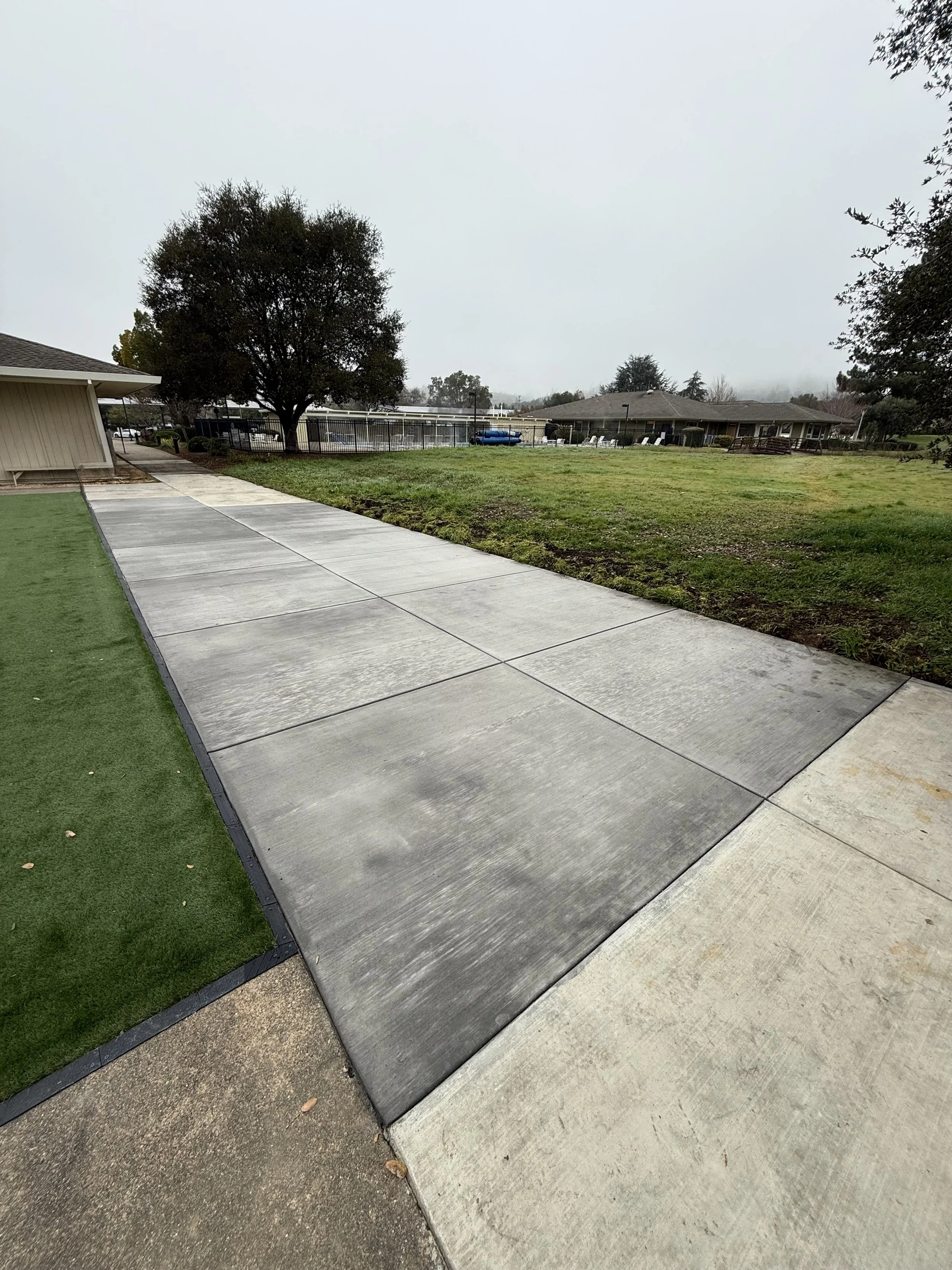 A sidewalk with concrete and asphalt sections next to a patch of artificial grass, surrounded by grass and trees, on a cloudy day at a residential area.