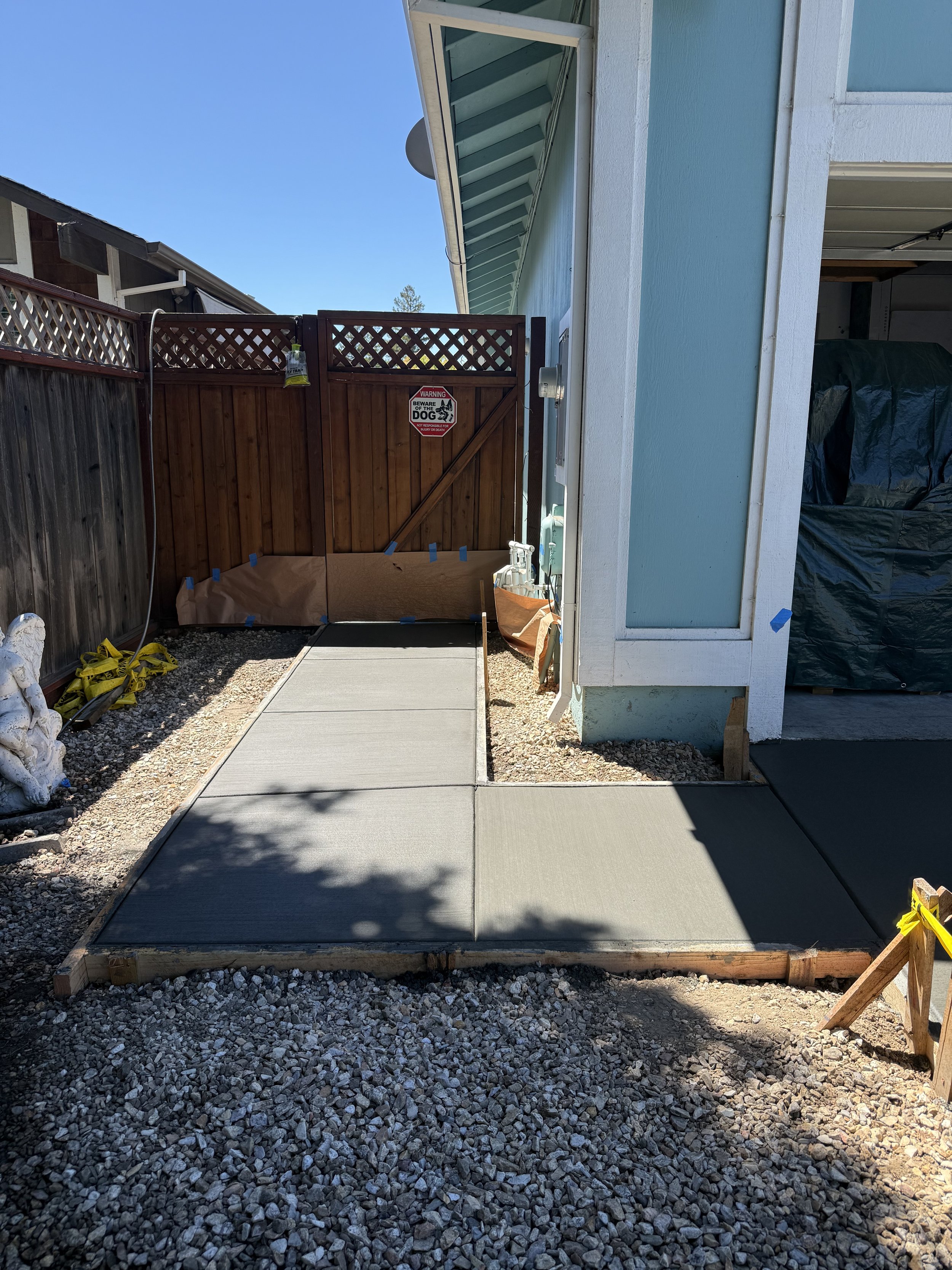 Concrete pathway leading to a wooden gate with a sign that says 'Warning: Beware of the Dog.' The pathway is bordered by gravel on both sides, and part of a house with light blue exterior walls is visible on the right.