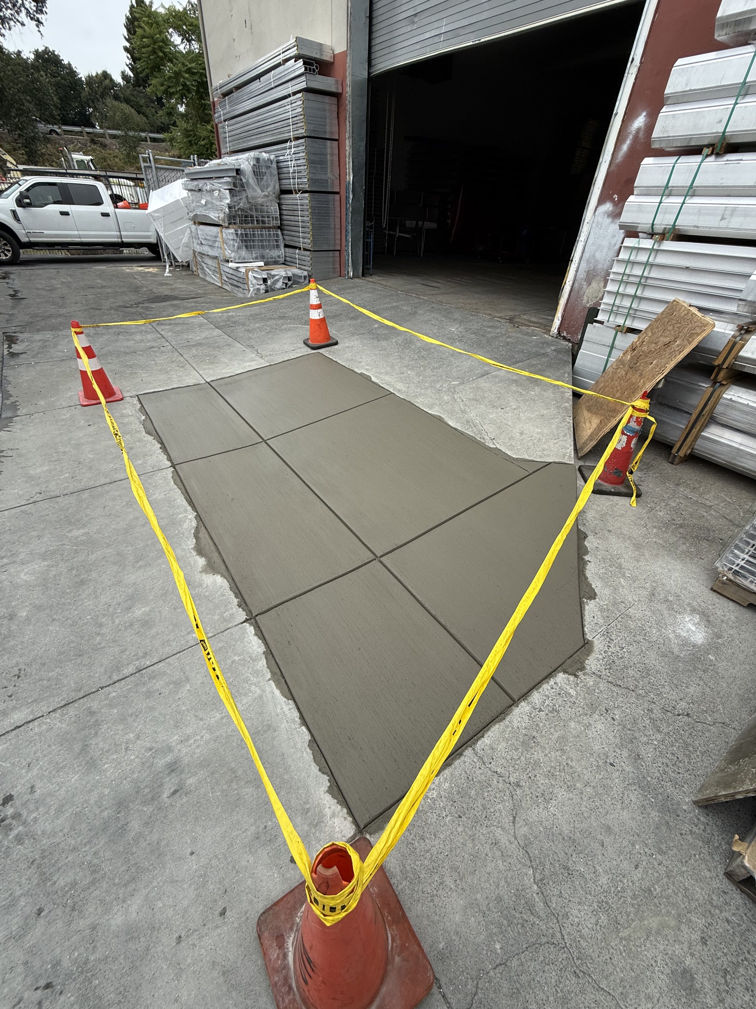 A sidewalk section being freshly poured with concrete, surrounded by yellow caution tape and orange traffic cones, outside a building with stored construction materials.