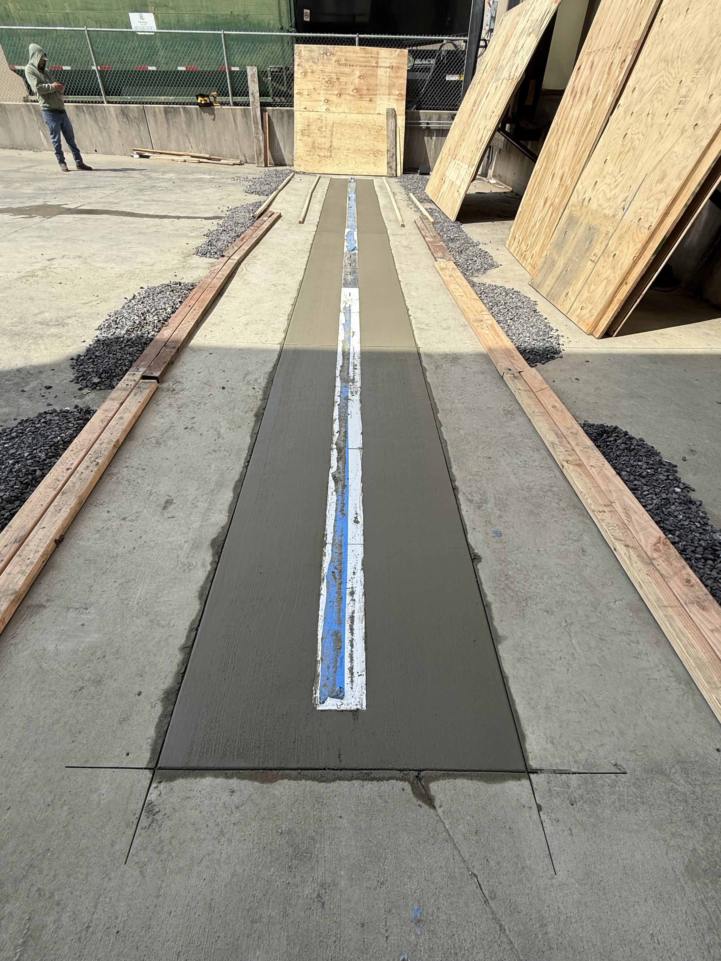 Construction site with fresh concrete being poured within wooden formwork, black gravel on edges, and plywood sheets leaning against the wall in the background; a person in a hoodie is standing and looking at a cell phone.