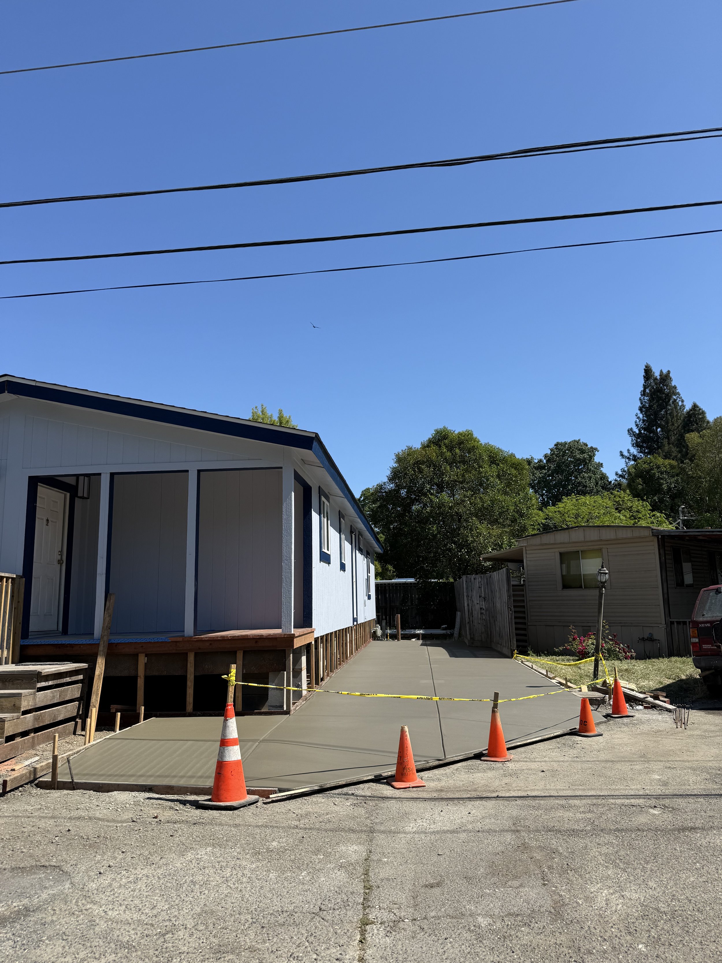 A sidewalk with orange cones and caution tape, leading to a house under construction with a newly poured concrete slab. In the background, there are trees and other houses, with power lines overhead and a clear blue sky.