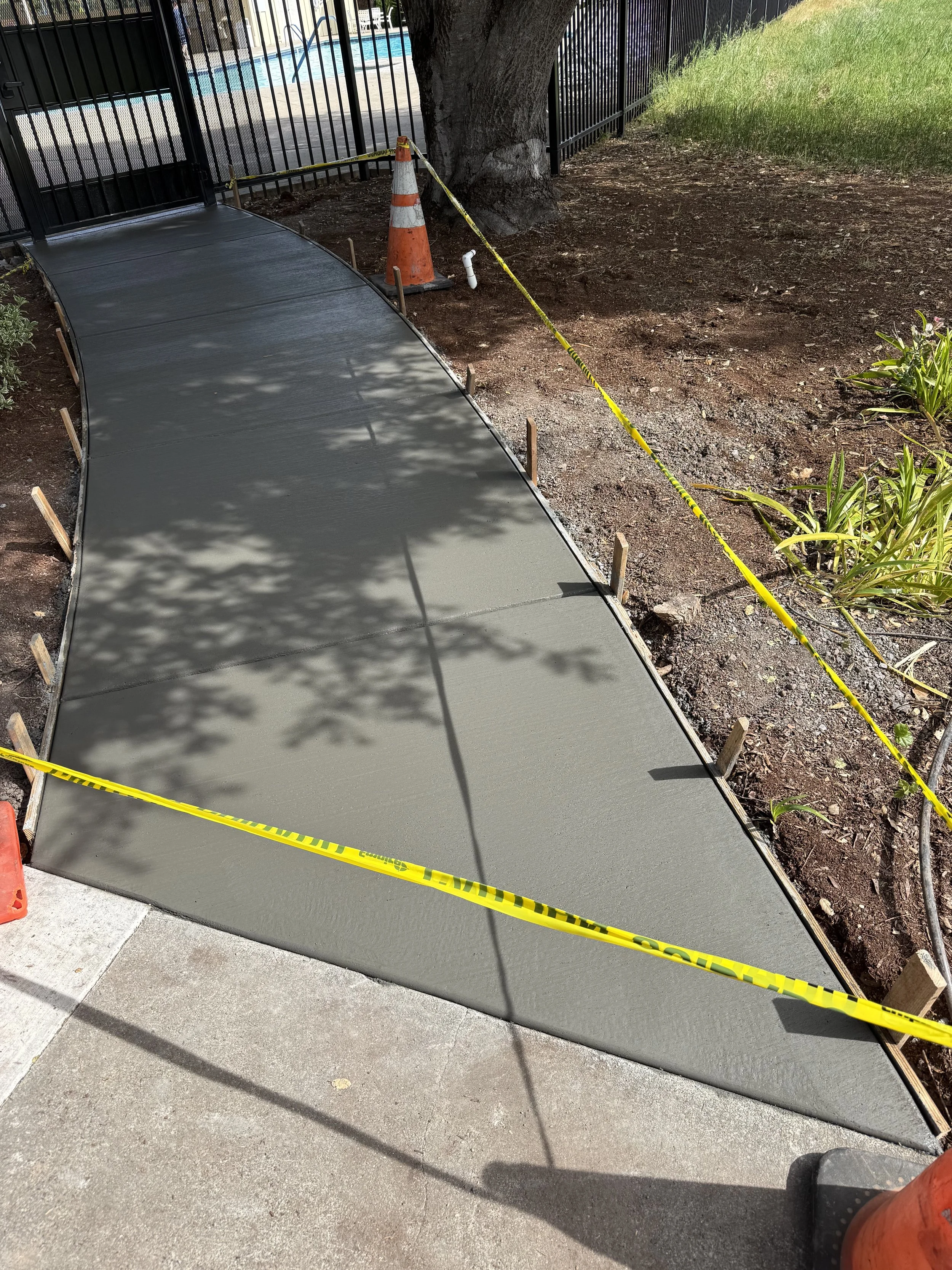 A freshly poured concrete sidewalk section being protected by yellow caution tape, with a traffic cone and orange construction marker nearby, under a large tree with a nearby grassy area and a black metal fence.