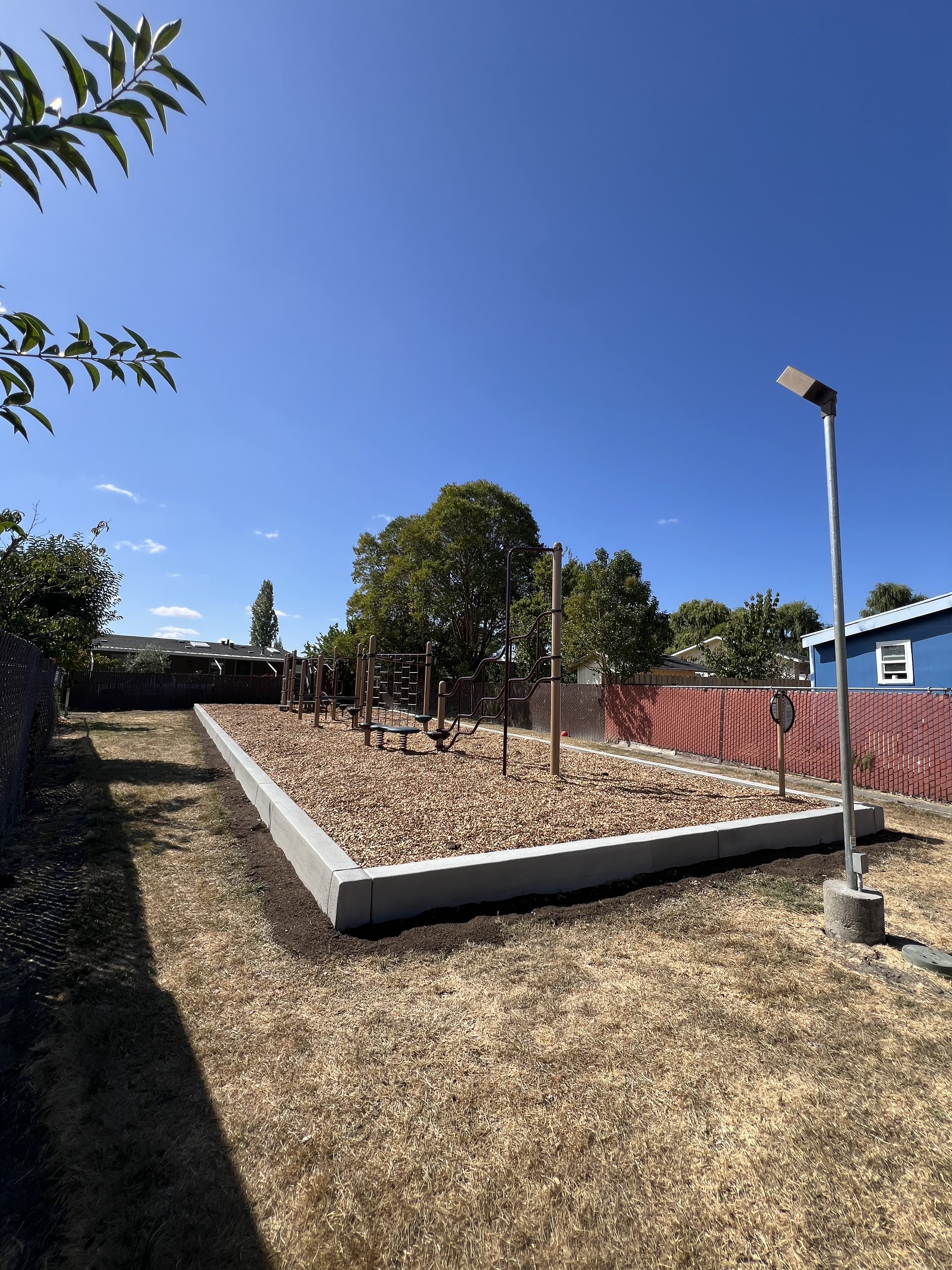 A playground area with outdoor workout equipment, a tall street light, trees, and a clear blue sky.