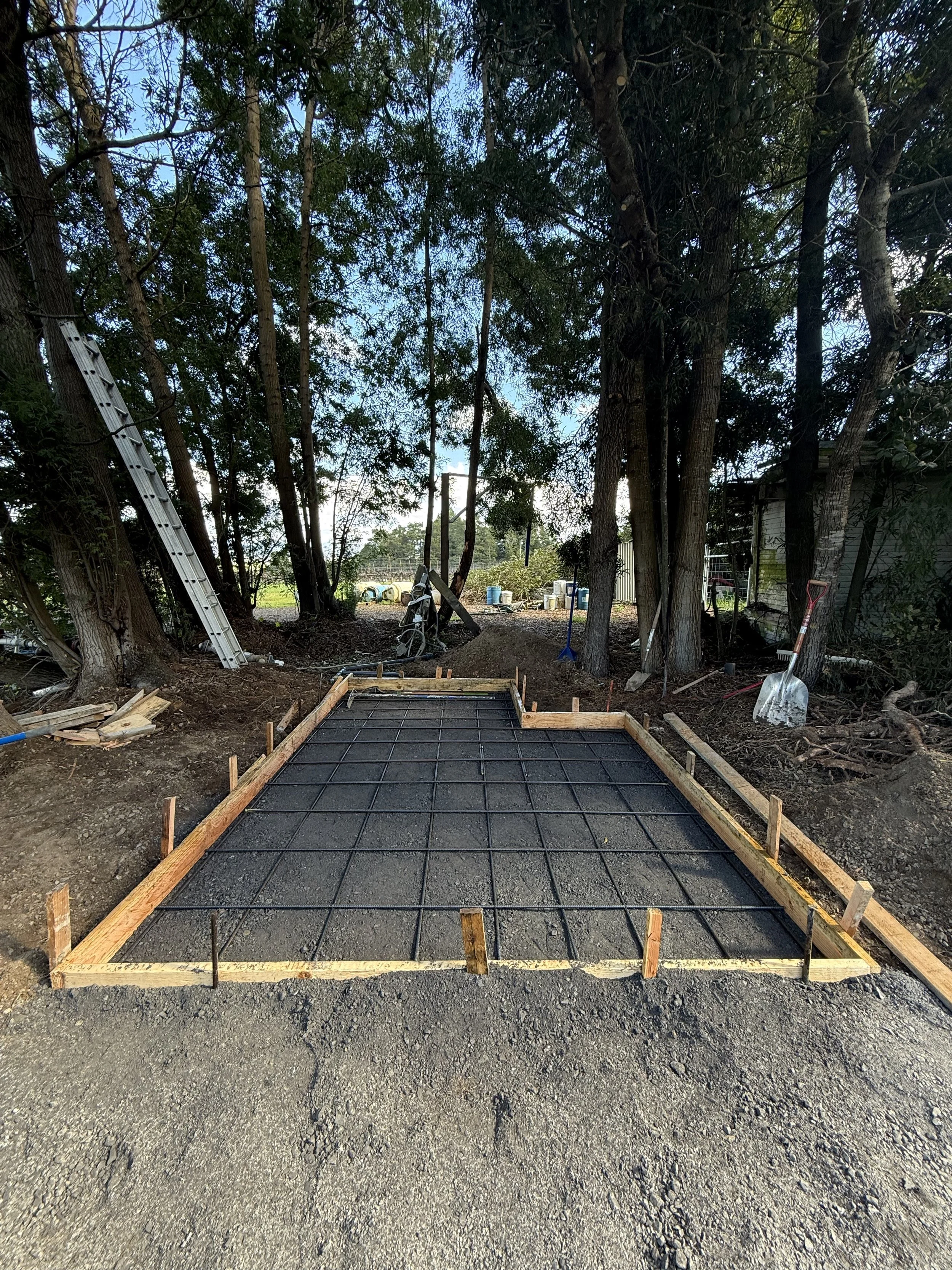 Construction site with a wooden frame for concrete foundation and a freshly poured concrete slab with reinforcement grid. Several trees and construction tools are visible in the background.