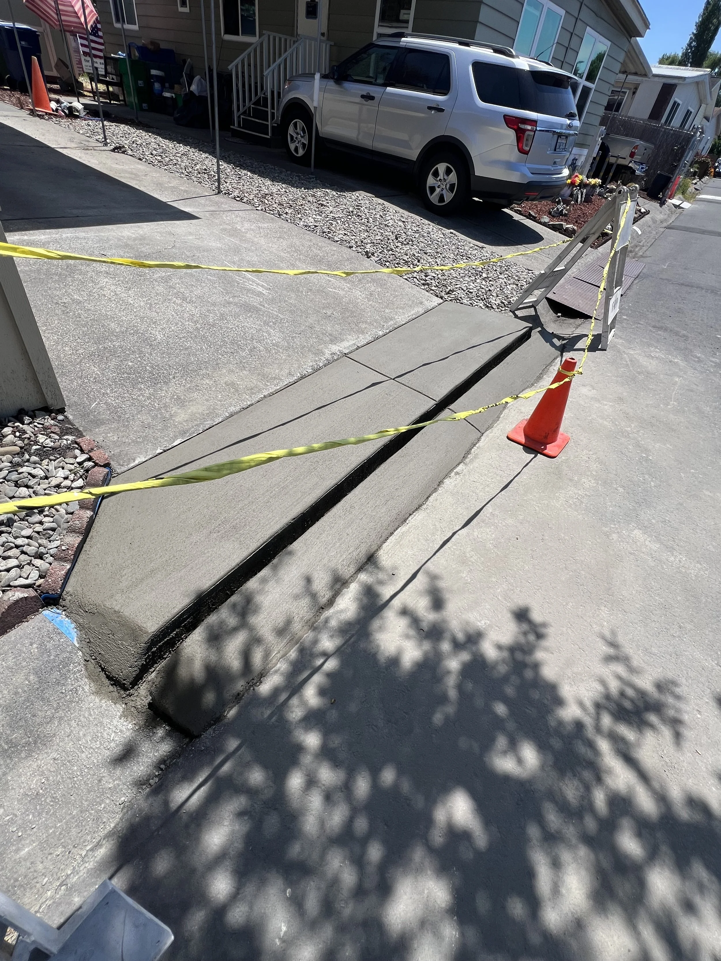 A construction or repair site outside a residential house with a sidewalk and driveway. There is caution tape and an orange traffic cone marking the area, and a large section of the sidewalk appears to be under construction or being replaced.