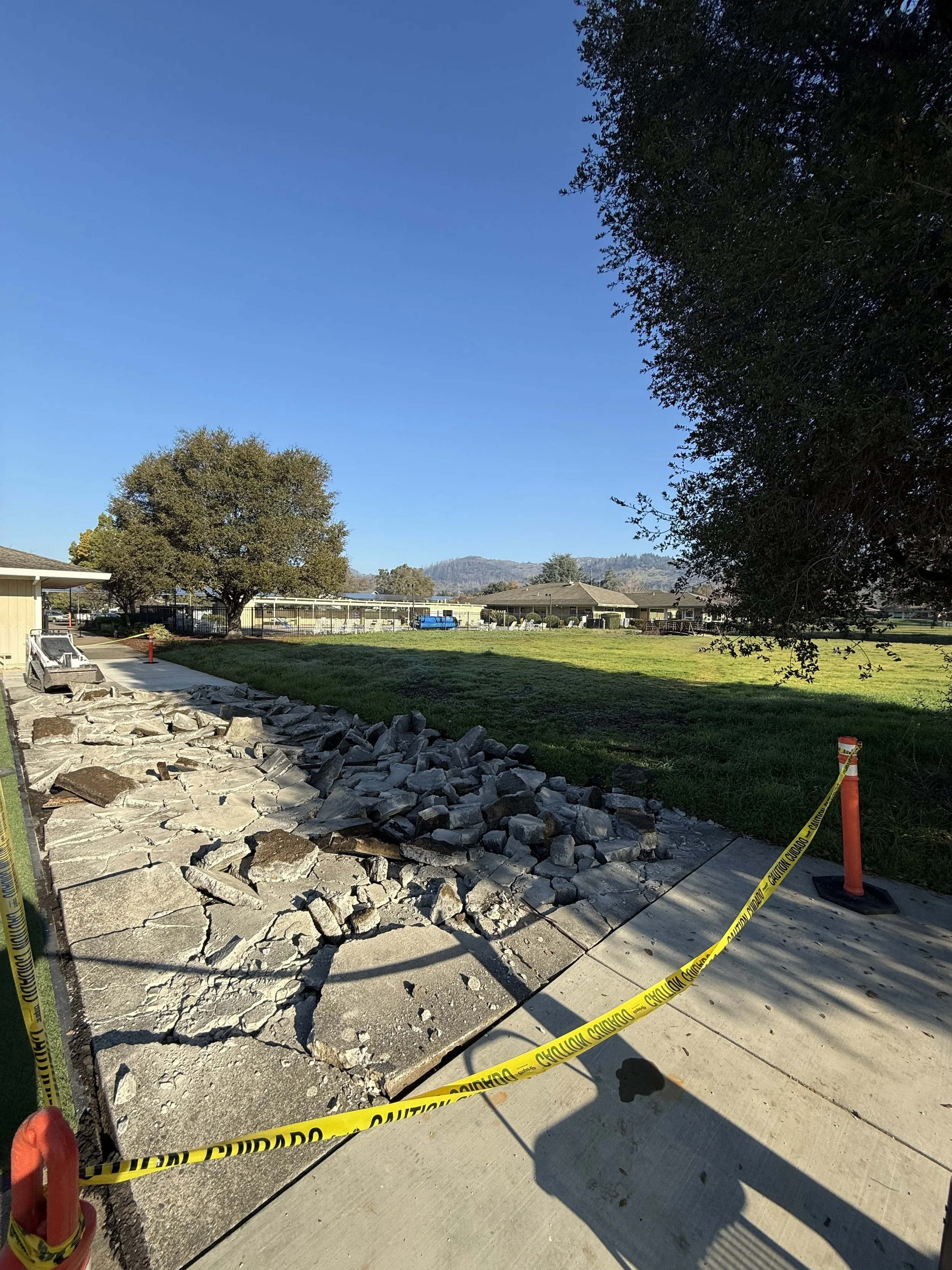 Sidewalk with damaged and cracked concrete, construction debris, caution tape, and cones, next to a grassy park with trees and buildings in the background.
