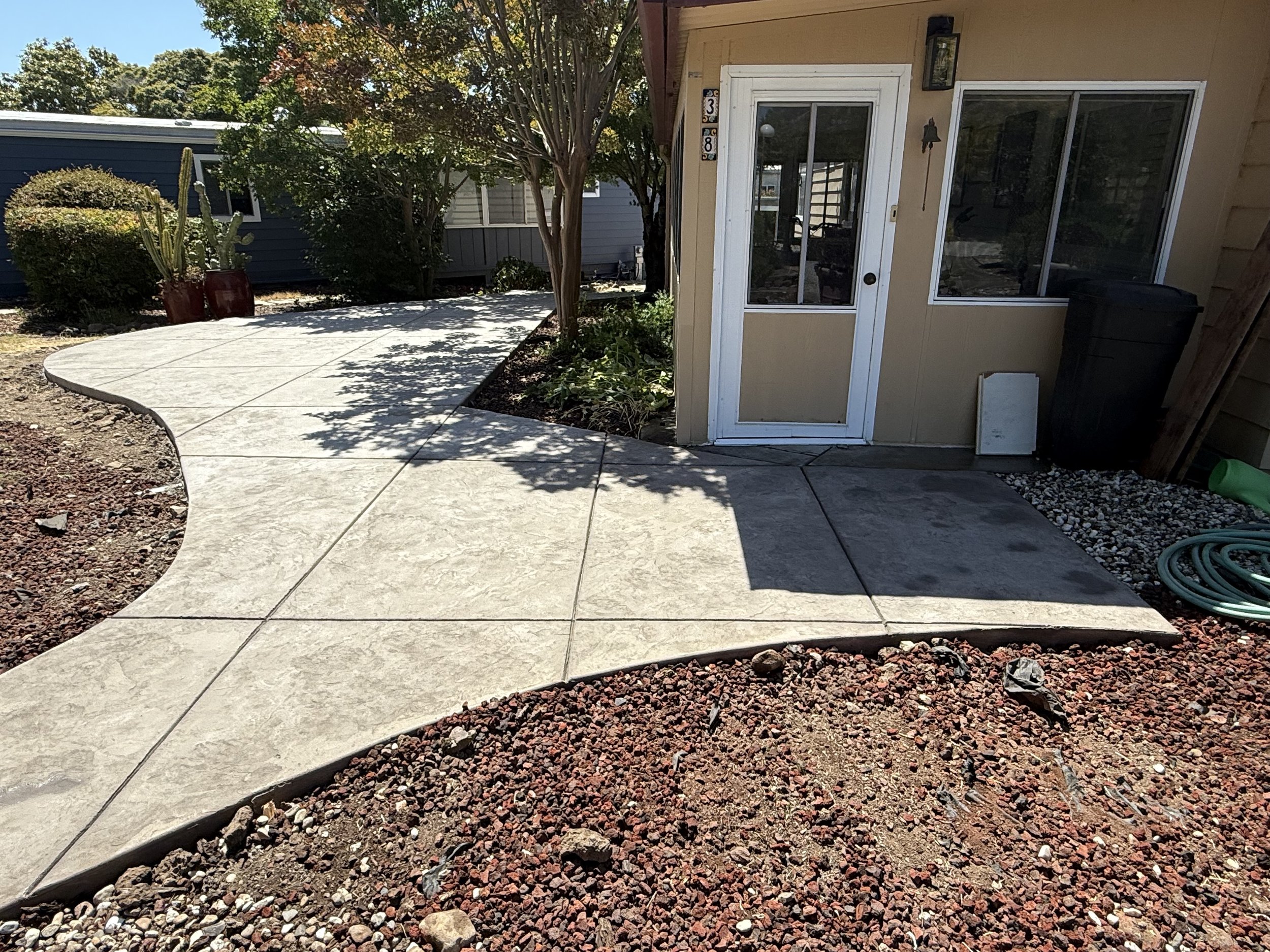 Backyard patio area with a concrete walkway, a small tree, bushes, and a house with large sliding glass door.