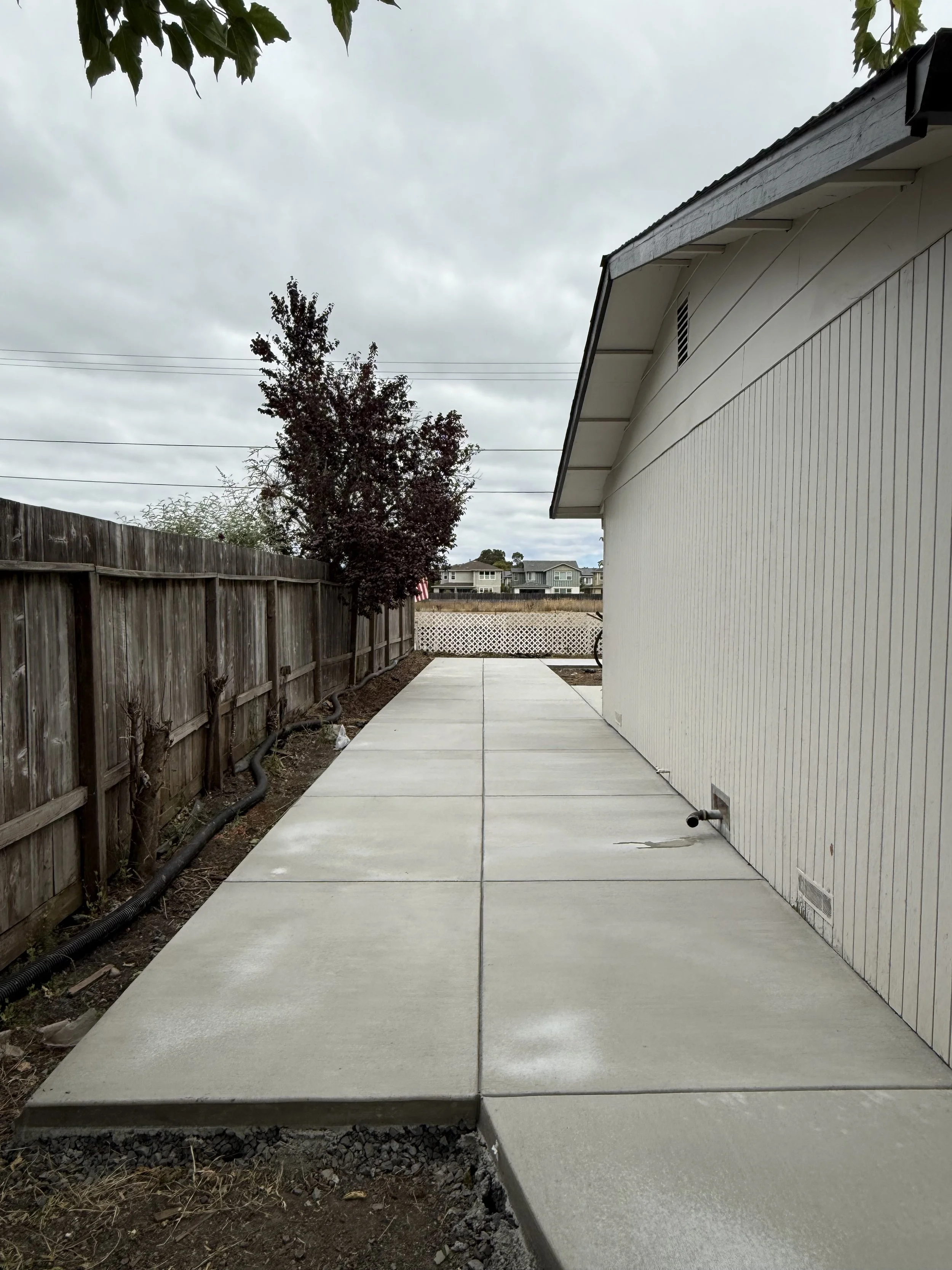 Newly poured concrete sidewalk next to a white house with wood siding, fenced backyard, and overcast sky.