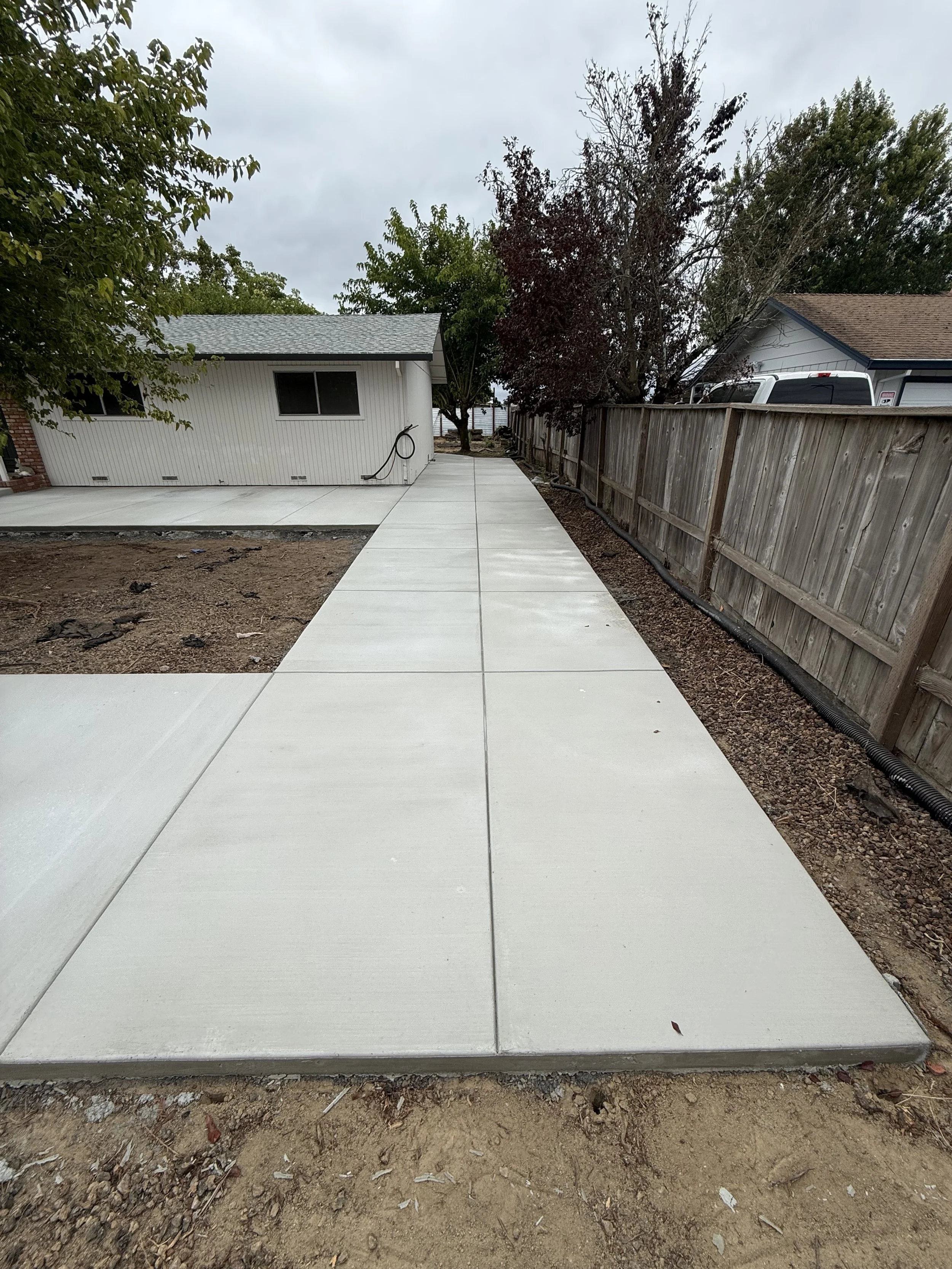 Newly poured concrete sidewalk next to a wooden fence, with a gray house and trees in the background on a cloudy day.