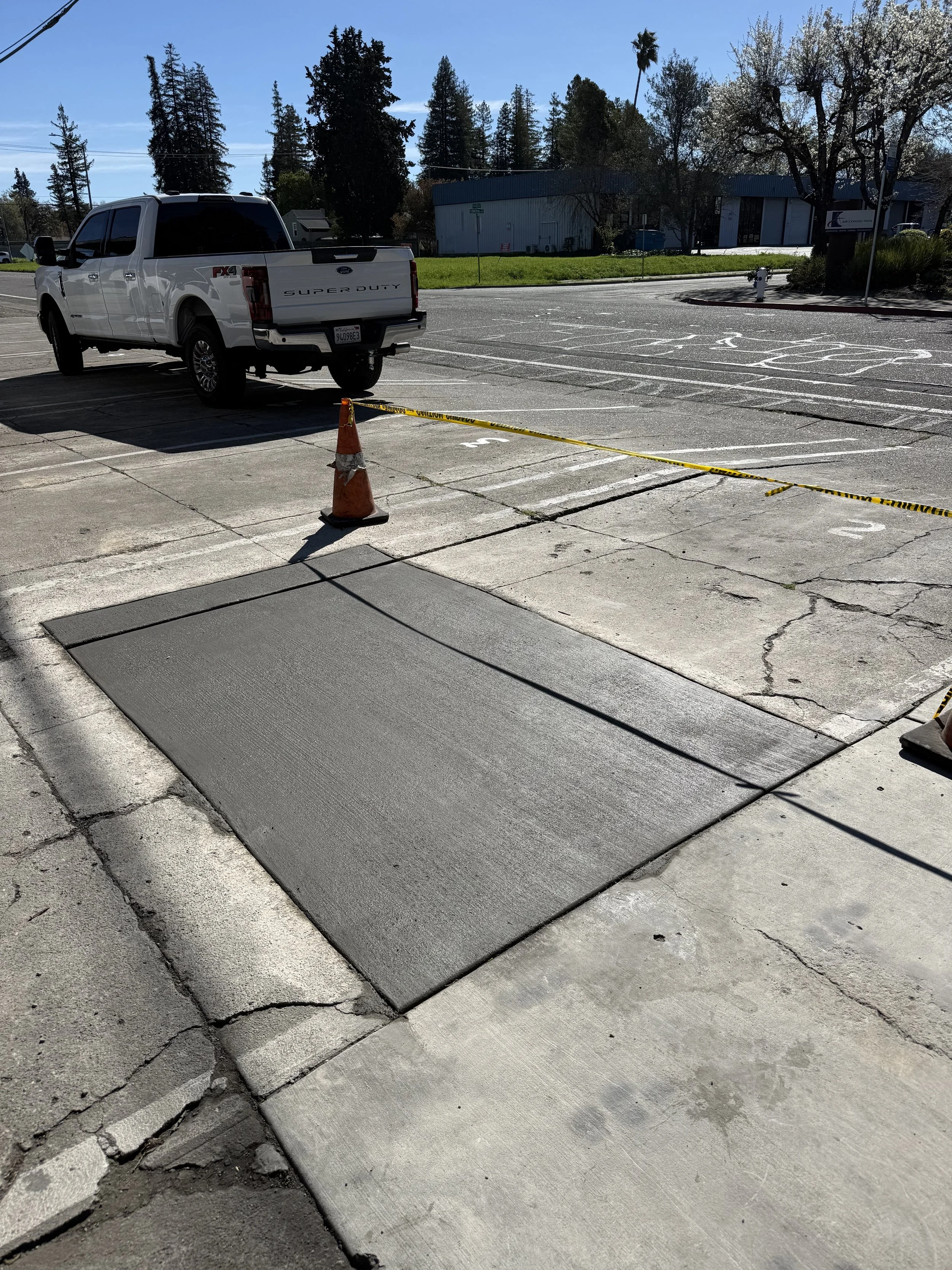 A partially damaged sidewalk with cracks and missing concrete sections, a traffic cone, a white pickup truck parked nearby, and a parking lot with painted lines.