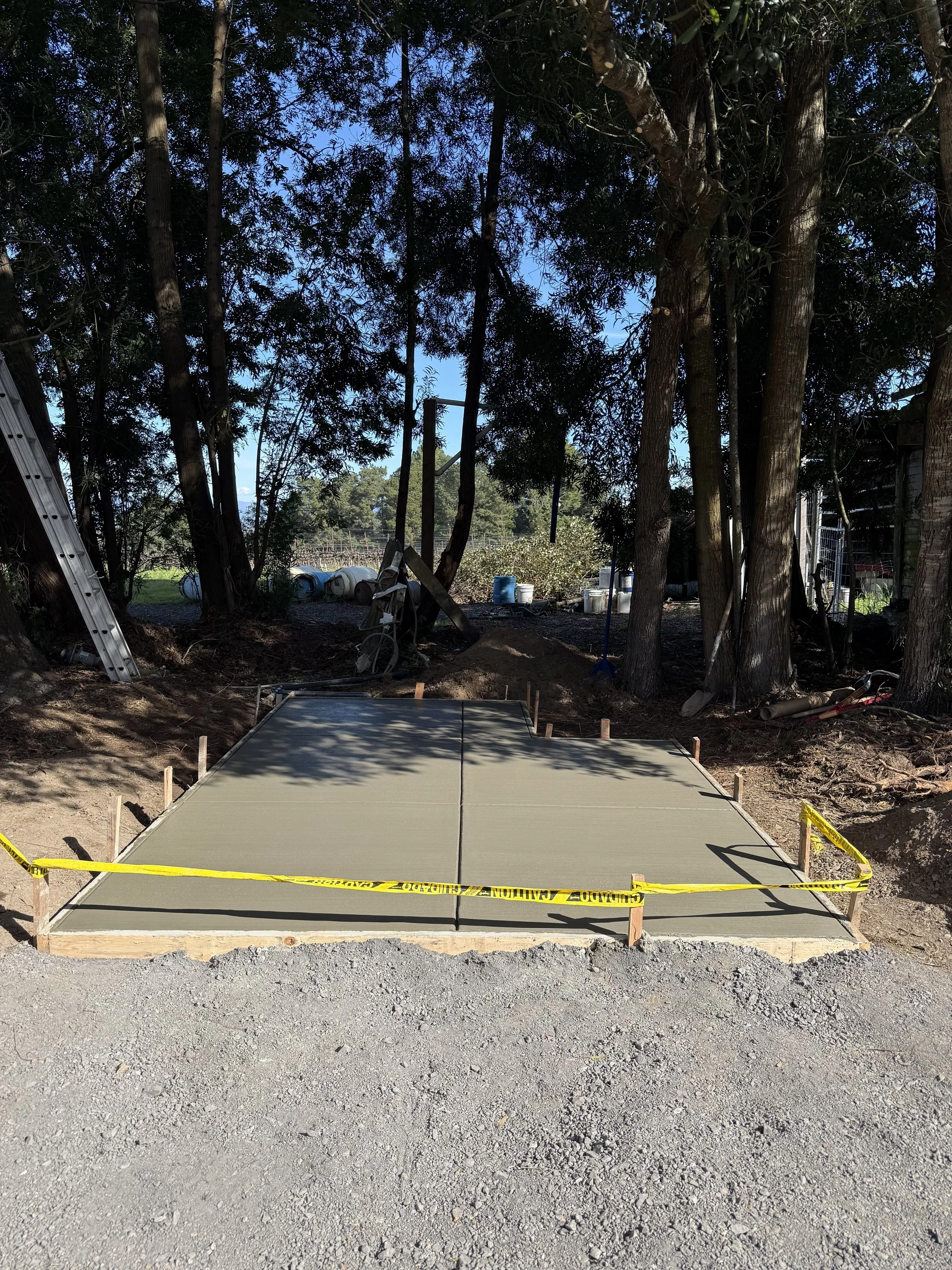 A freshly poured concrete slab on a construction site, surrounded by wooden forms and yellow caution tape, with trees and a blue sky in the background.